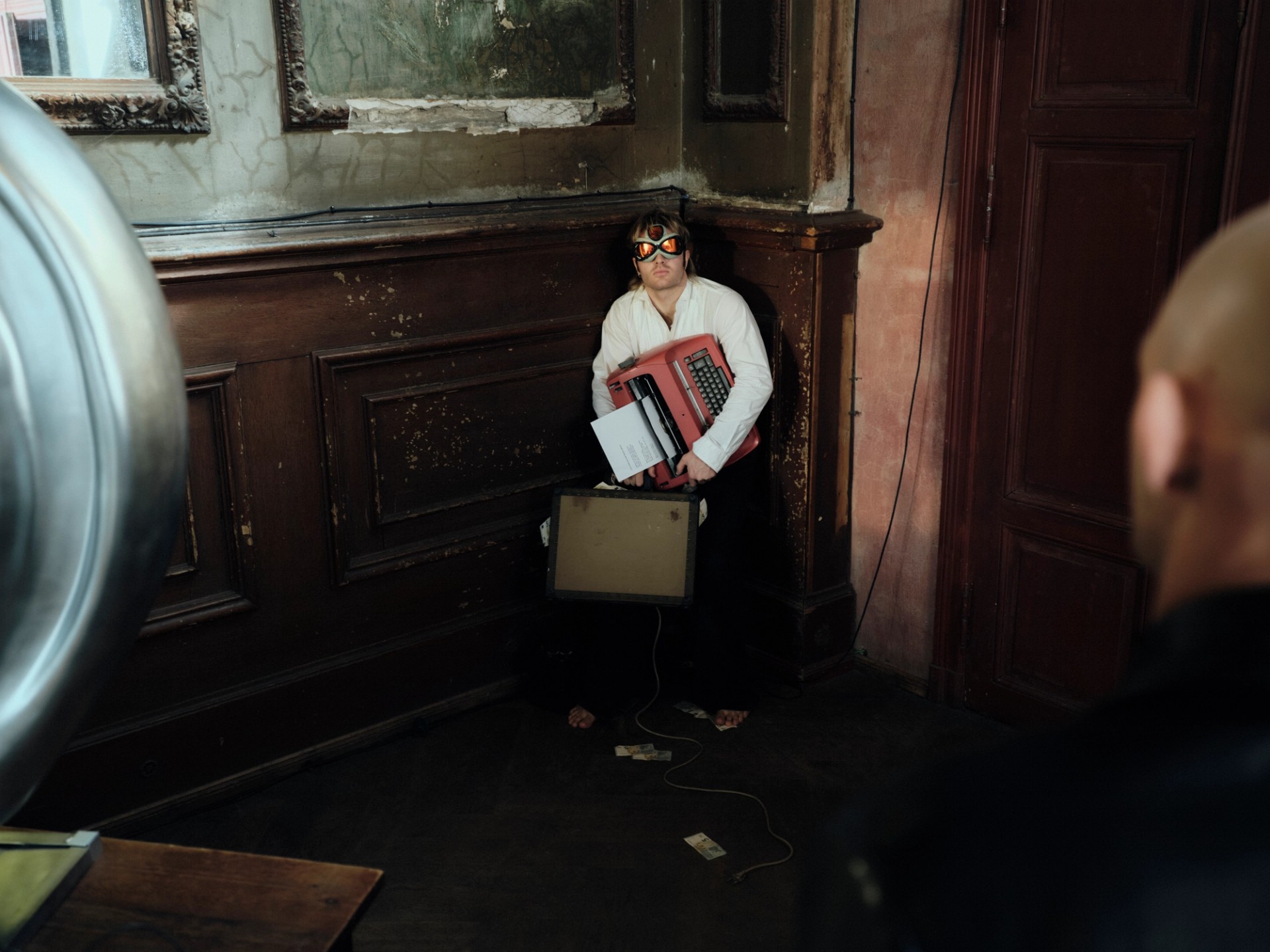 A performer holding an accordion stands beside a wood-panelled wall and a large metallic ear-shaped speaker.