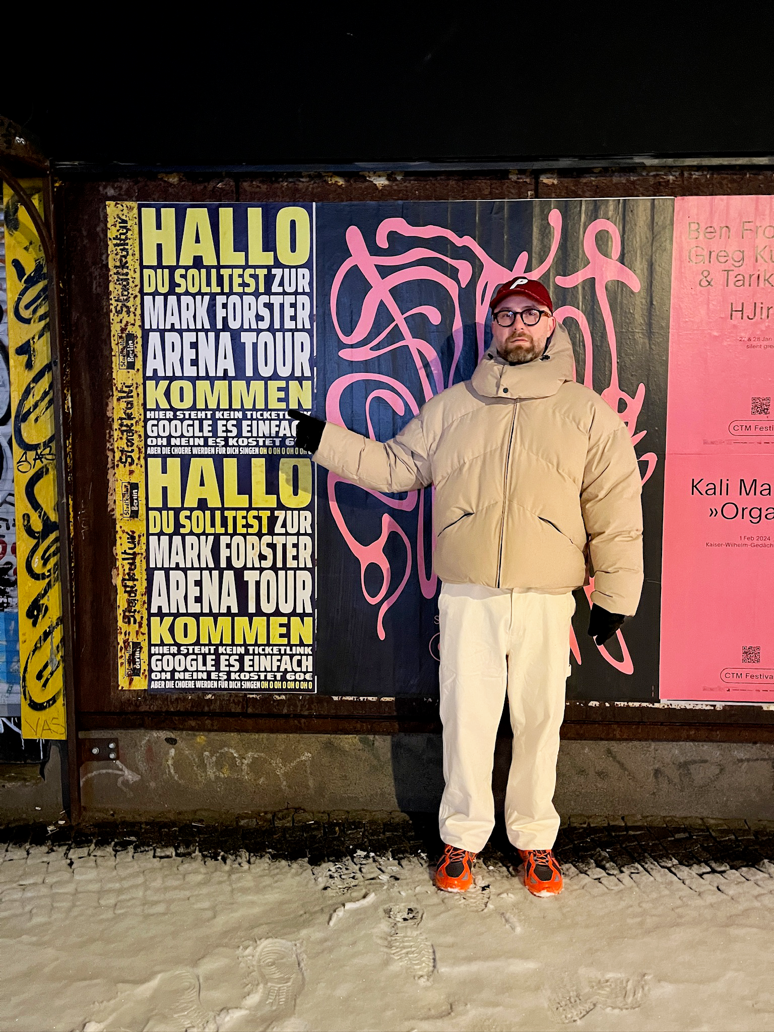 Mark Forster in a beige puffer jacket and red cap stands in the snow at night, pointing at the poster "HALLO DU SOLLTEST ZUR MARK FORSTER ARENA TOUR KOMMEN".