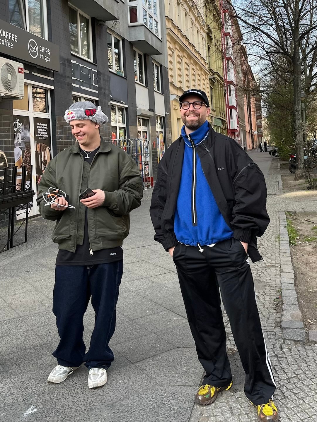 On a Berlin pavement, Papaplatte looks at his phone in a grey trapper hat and green bomber jacket while Mark Forster laughs in a black track jacket over a blue fleece.