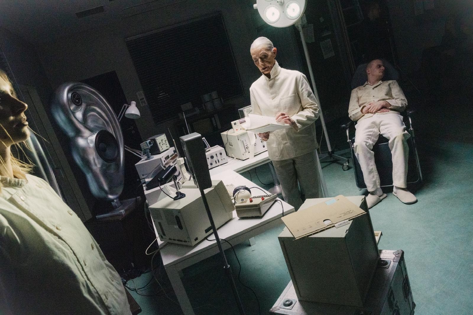 Tilted view of the SkiAgguMusik clinical set, with white-clad figures, a desk and a metallic ear-shaped speaker.