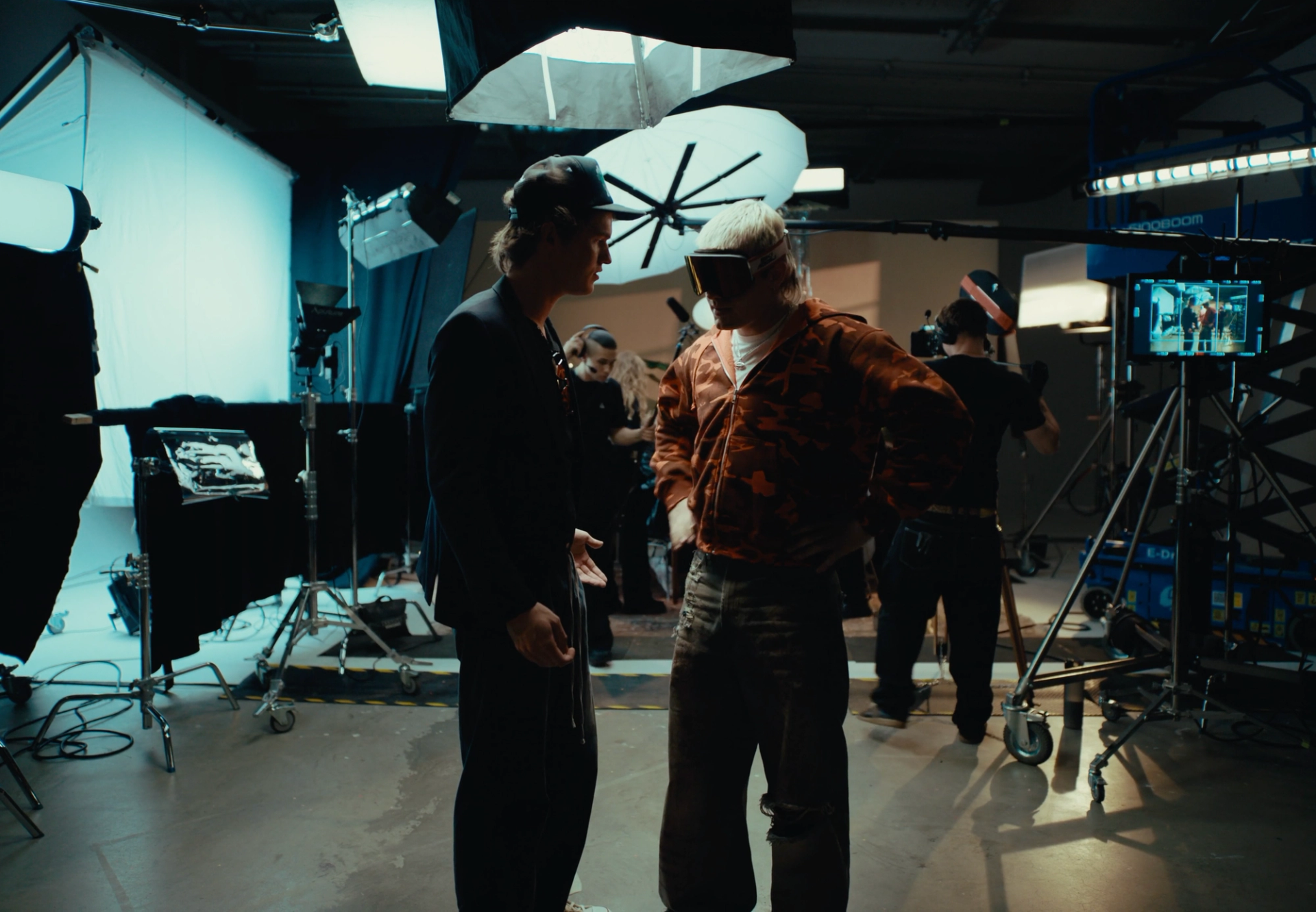 Film studio with softboxes, light stands and a monitor: Flaschko in a cap and Ski Aggu in goggles stand talking while crew work behind them.