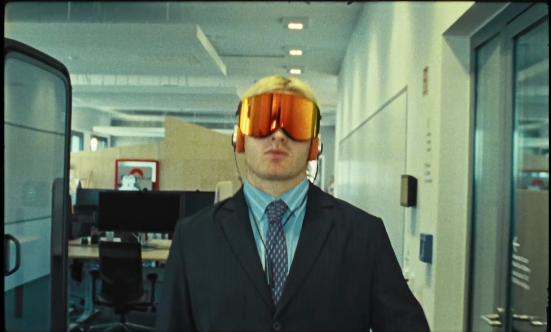 Full frame of Ski Aggu walking down an office corridor in orange ski goggles and headphones, desks and monitors behind him.