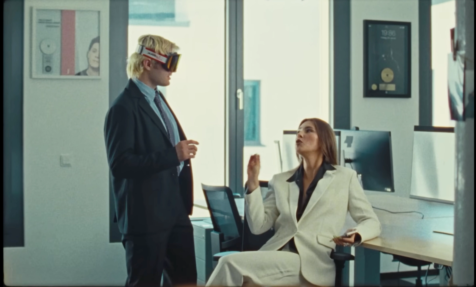 Trailer still: Ski Aggu in suit and ski goggles stands beside Stefanie Giesinger, who sits at an office desk in a white suit, gesturing as she talks.
