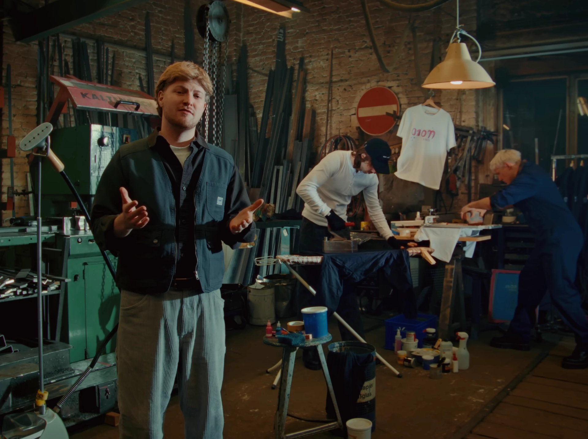 A band member talks to the camera in a metal workshop; behind him another works at a table next to a craftsman in blue overalls.