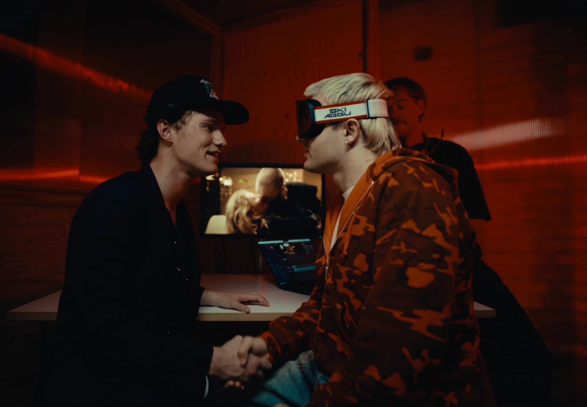Handshake in a red-lit room: Flaschko in a cap and Ski Aggu in goggles face each other in front of an edit monitor, Aditotoro behind them.