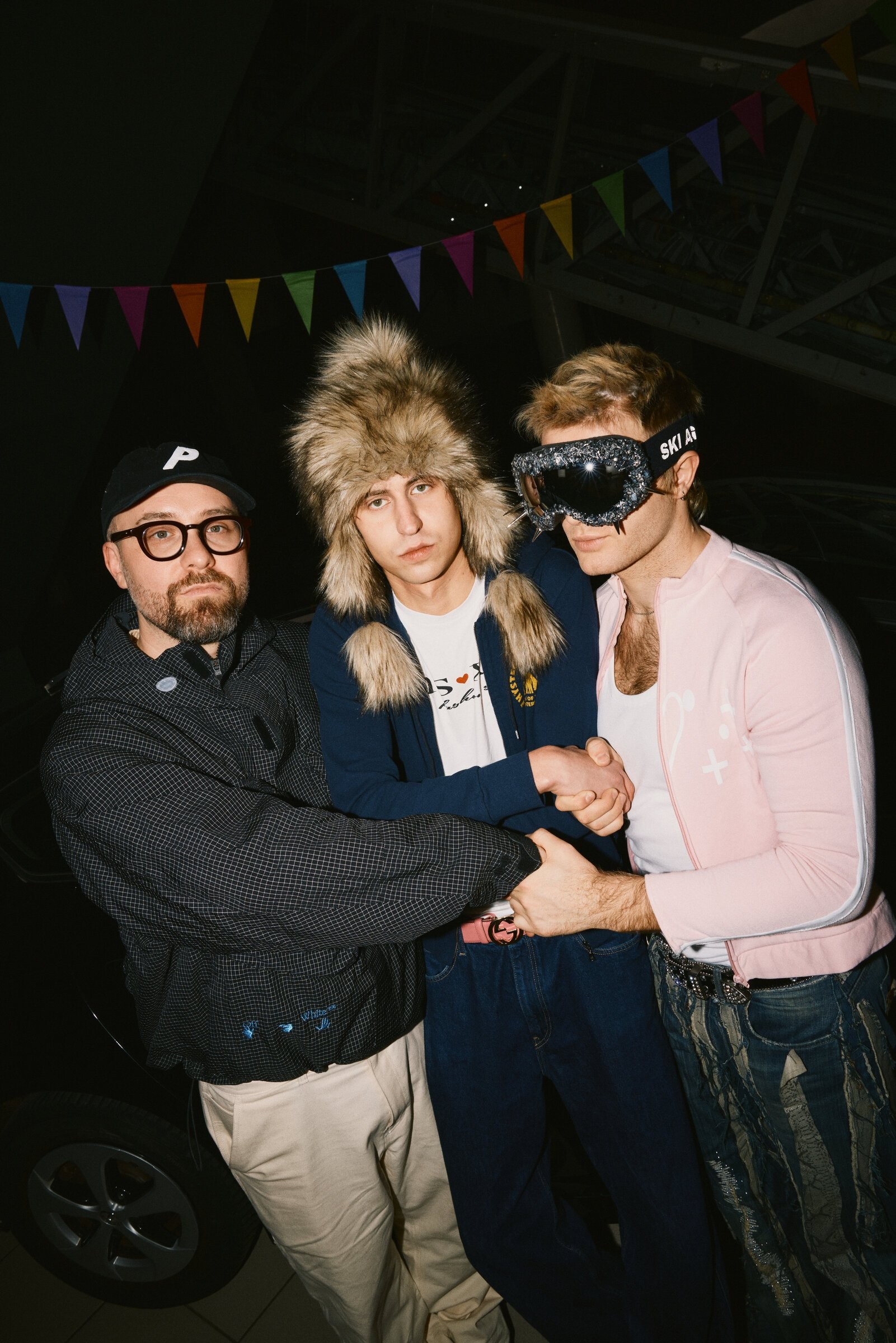 Flash photo under coloured bunting: Mark Forster in a black cap and checked jacket, Skrt Cobain in a large fur hat and Ski Aggu in glittering ski goggles and a pink jacket.