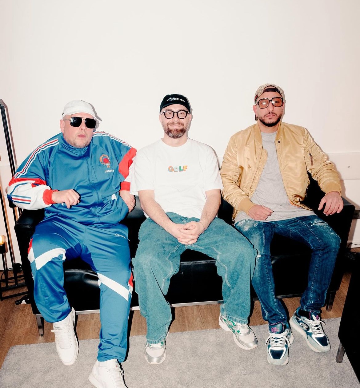 Three men on a black leather sofa: Mark Forster in a white "GOLF" T-shirt and wide jeans between Celo in a blue retro tracksuit and Abdi in a gold bomber jacket.