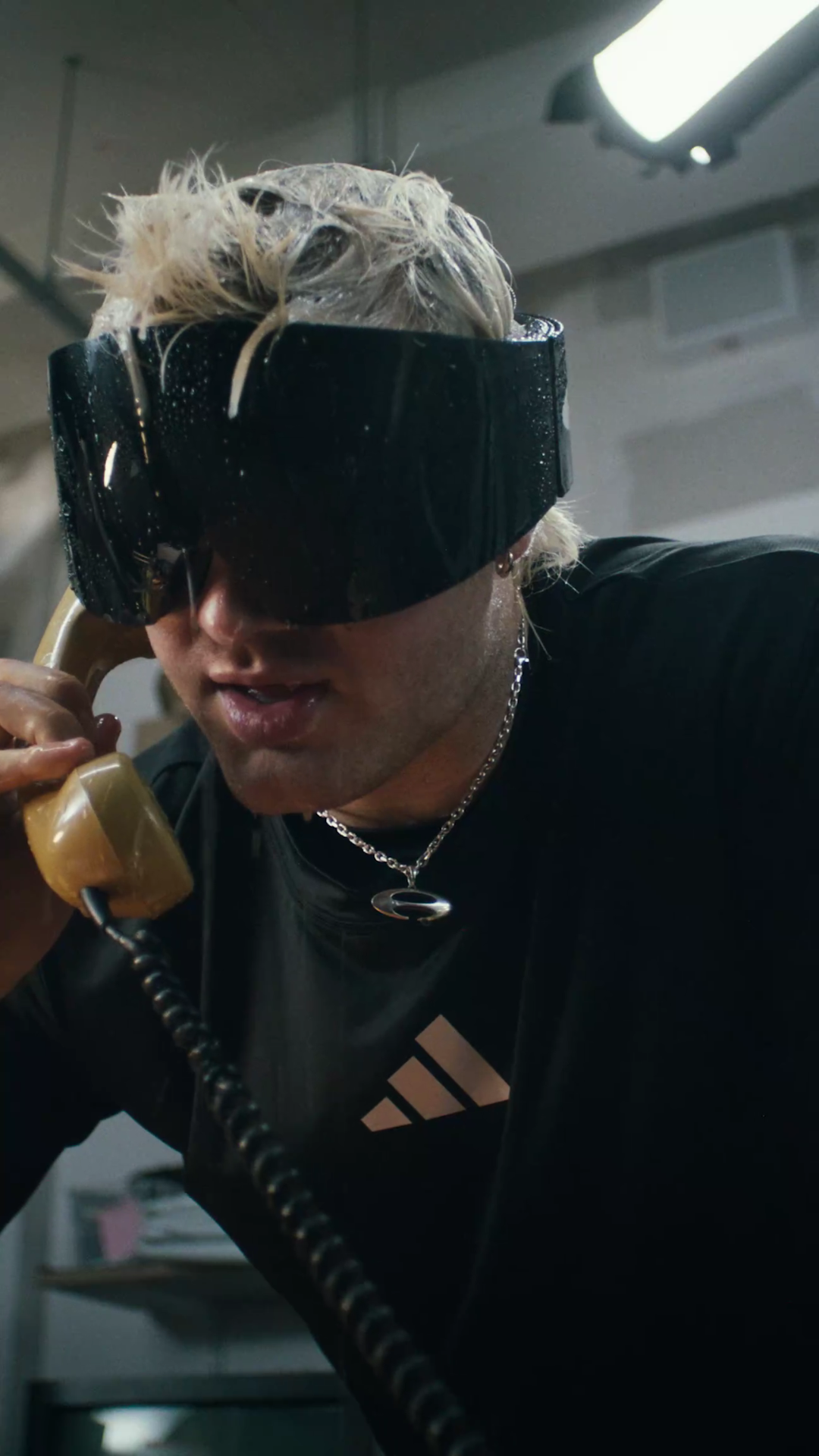 Low-angle close-up of Ski Aggu in black wraparound ski goggles and a black Adidas shirt, speaking into a yellow phone receiver with a coiled cord.