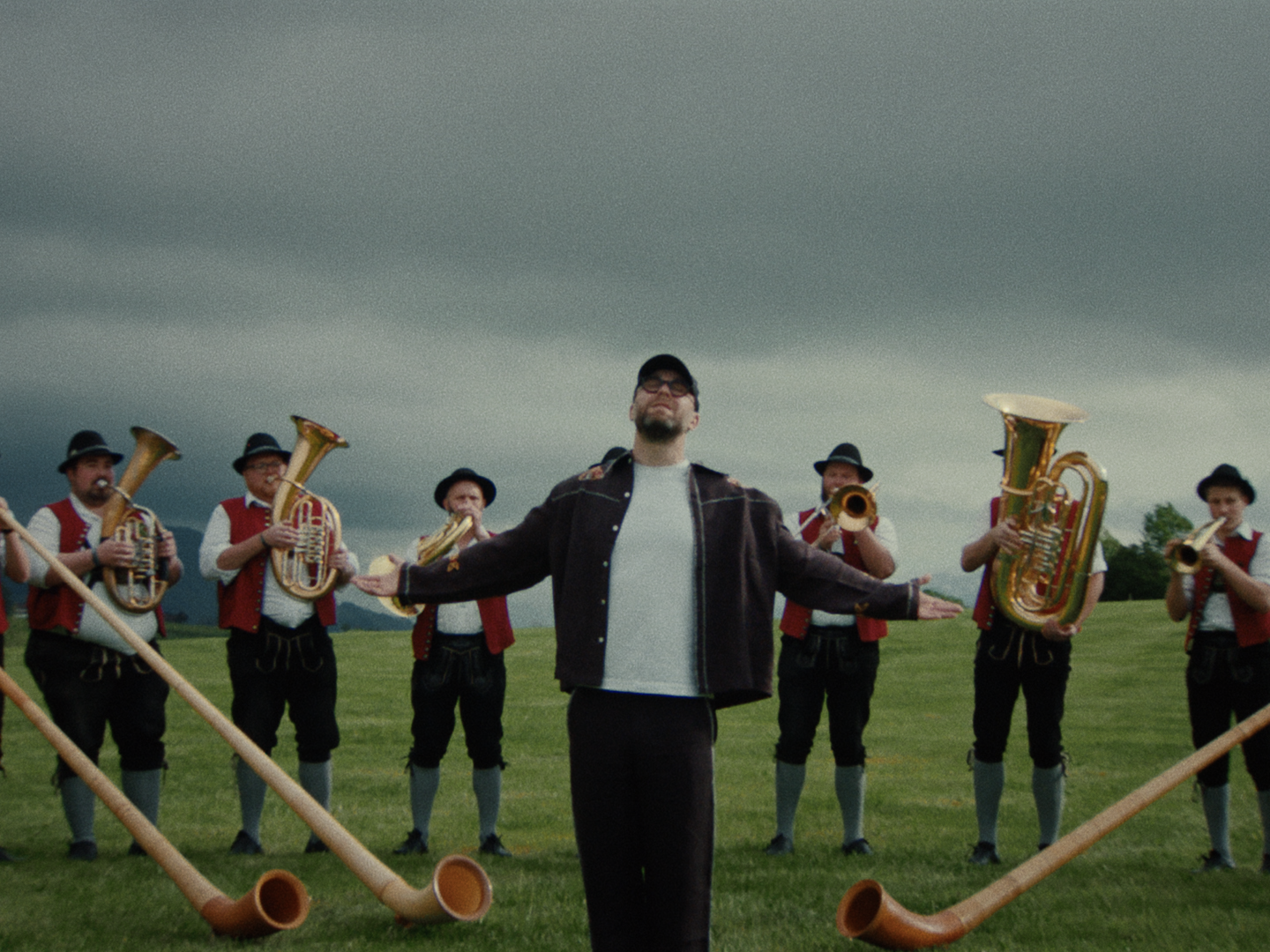 Low-angle view of Mark Forster with eyes closed and arms spread under a dark sky, brass players and alphorns lined up behind him.