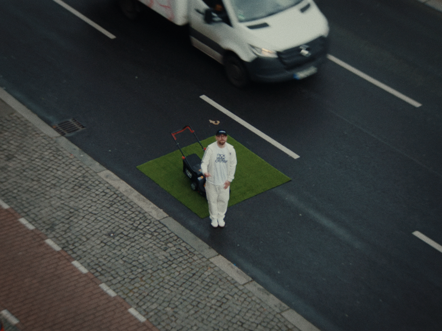 Seen from above, Mark Forster in white stands on a square of turf with a lawn mower in the middle of a city street as a van drives past.
