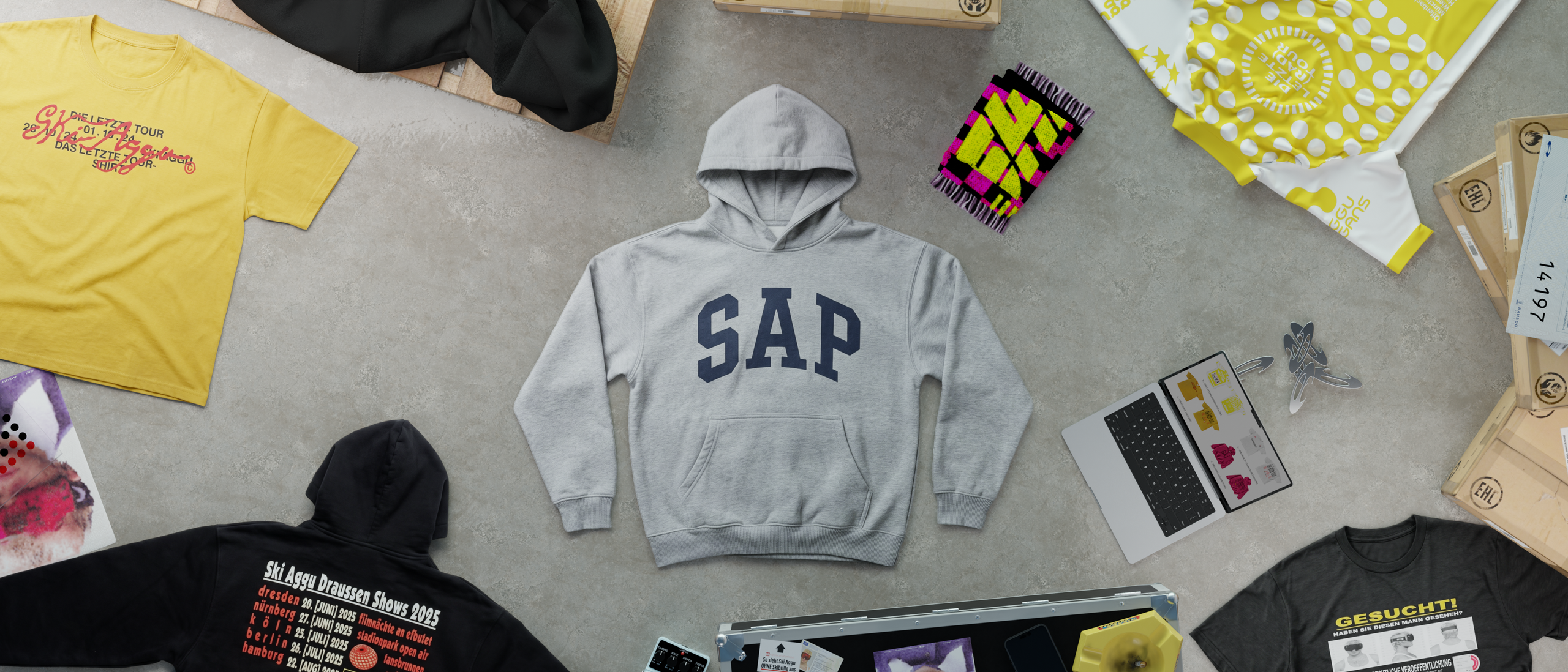 Merch laid out around the grey hoodie, its arms now hanging down, with the yellow tour T-shirt at top left and the black wanted-poster shirt at bottom right.