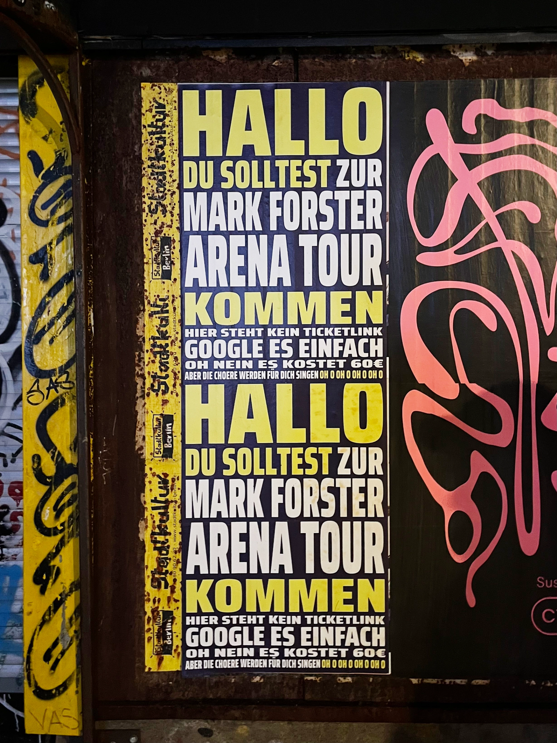 Text-only tour poster in yellow and white capitals on dark blue, pasted twice one above the other on a rusty wall next to graffiti.