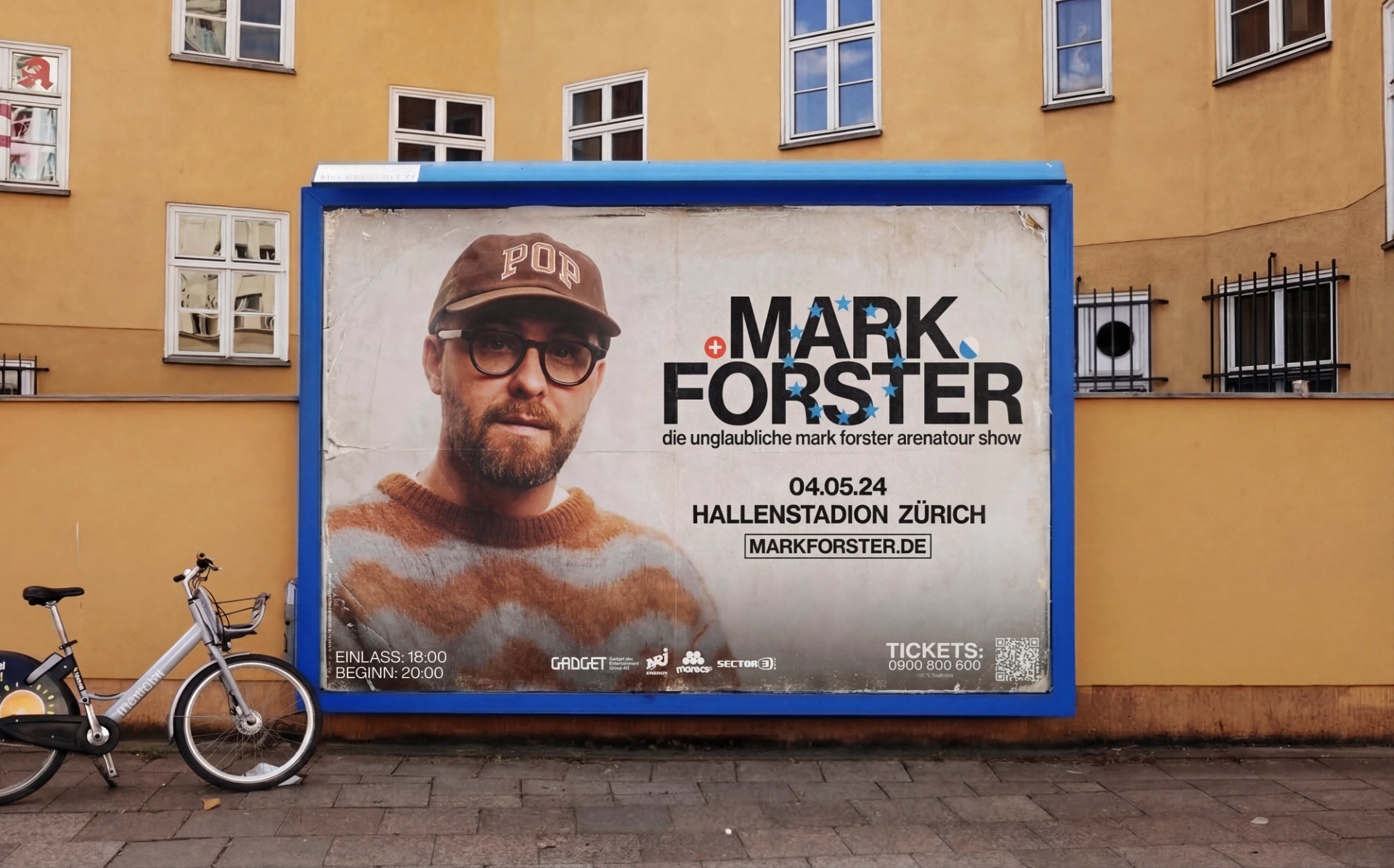 Billboard on a yellow house wall: the artist in a brown cap and striped jumper beside "MARK FORSTER", announcing the Hallenstadion Zürich show on 04.05.24.