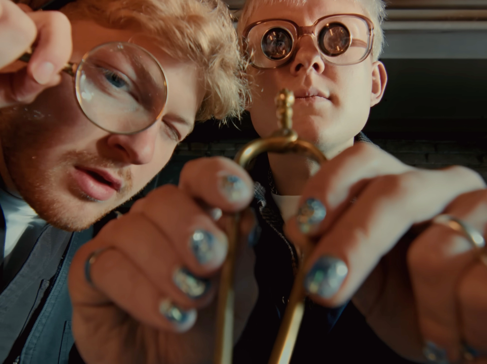 Taller crop of the inspection scene: glittery blue nail polish on the hands holding the brass part, the magnifying glass enlarging one eye.