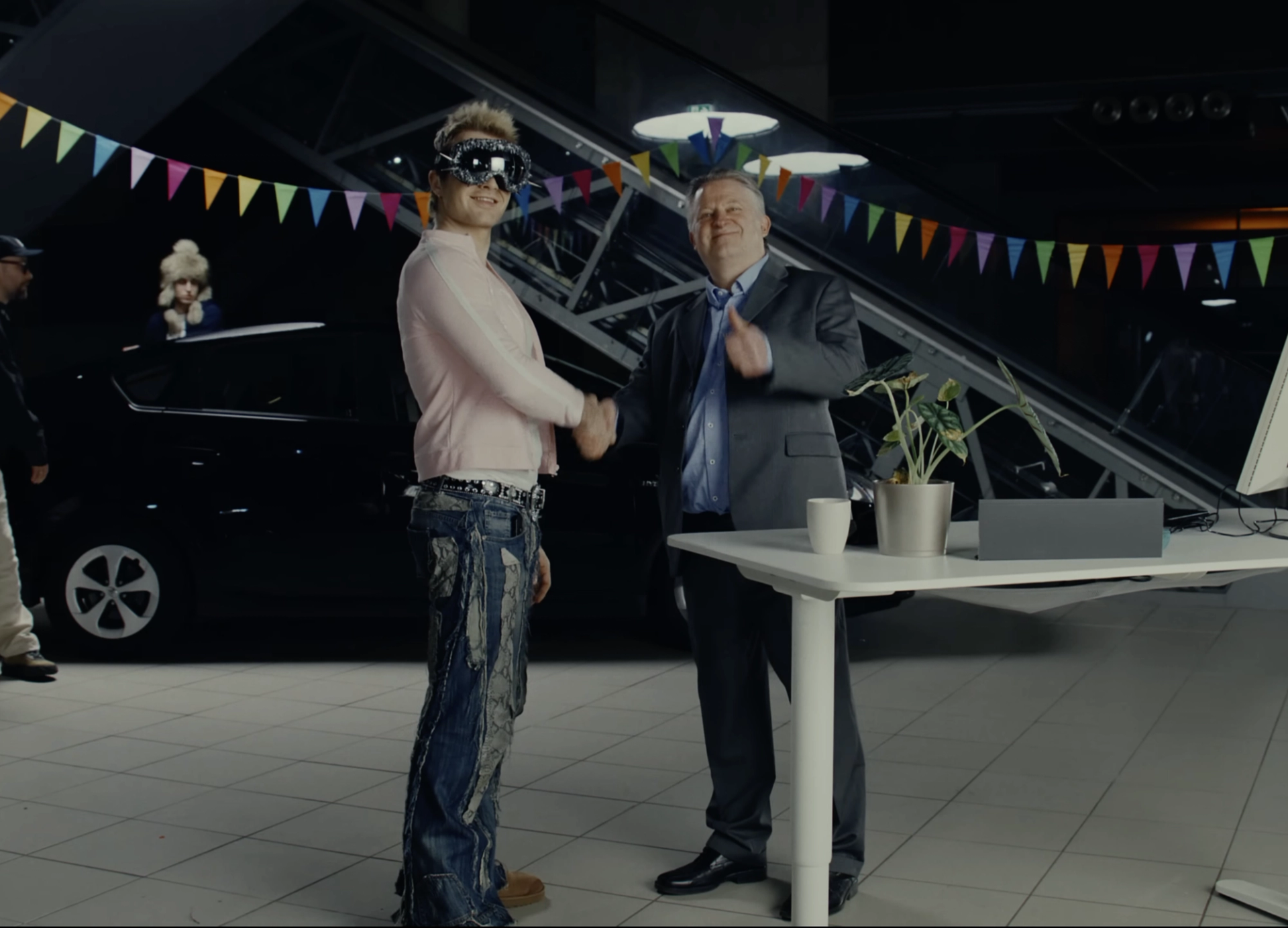 Car showroom hung with colourful bunting: Ski Aggu in studded goggles, a pink shirt and patched jeans shakes hands with a salesman in a grey suit.
