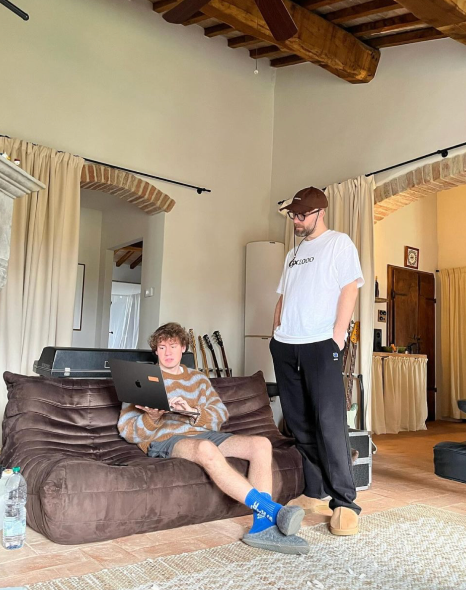Farmhouse living room with wooden beams: Mark Forster in a white T-shirt and brown cap stands beside Nick Ulrich, who works on a laptop on a brown sofa, guitars lined up behind.