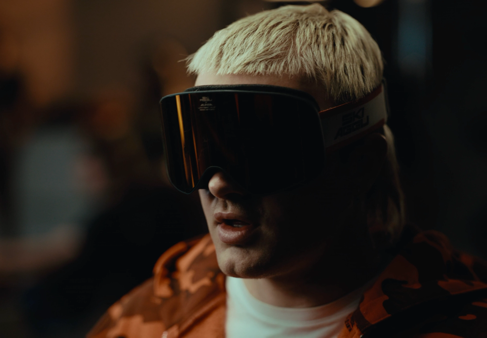 Tight portrait of Ski Aggu in dark ski goggles, bleached hair above and the collar of the orange camouflage hoodie below.