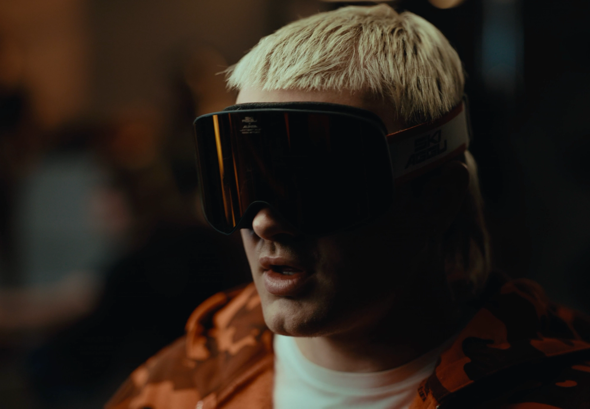 Tight portrait of Ski Aggu in dark ski goggles, bleached hair above and the collar of the orange camouflage hoodie below.