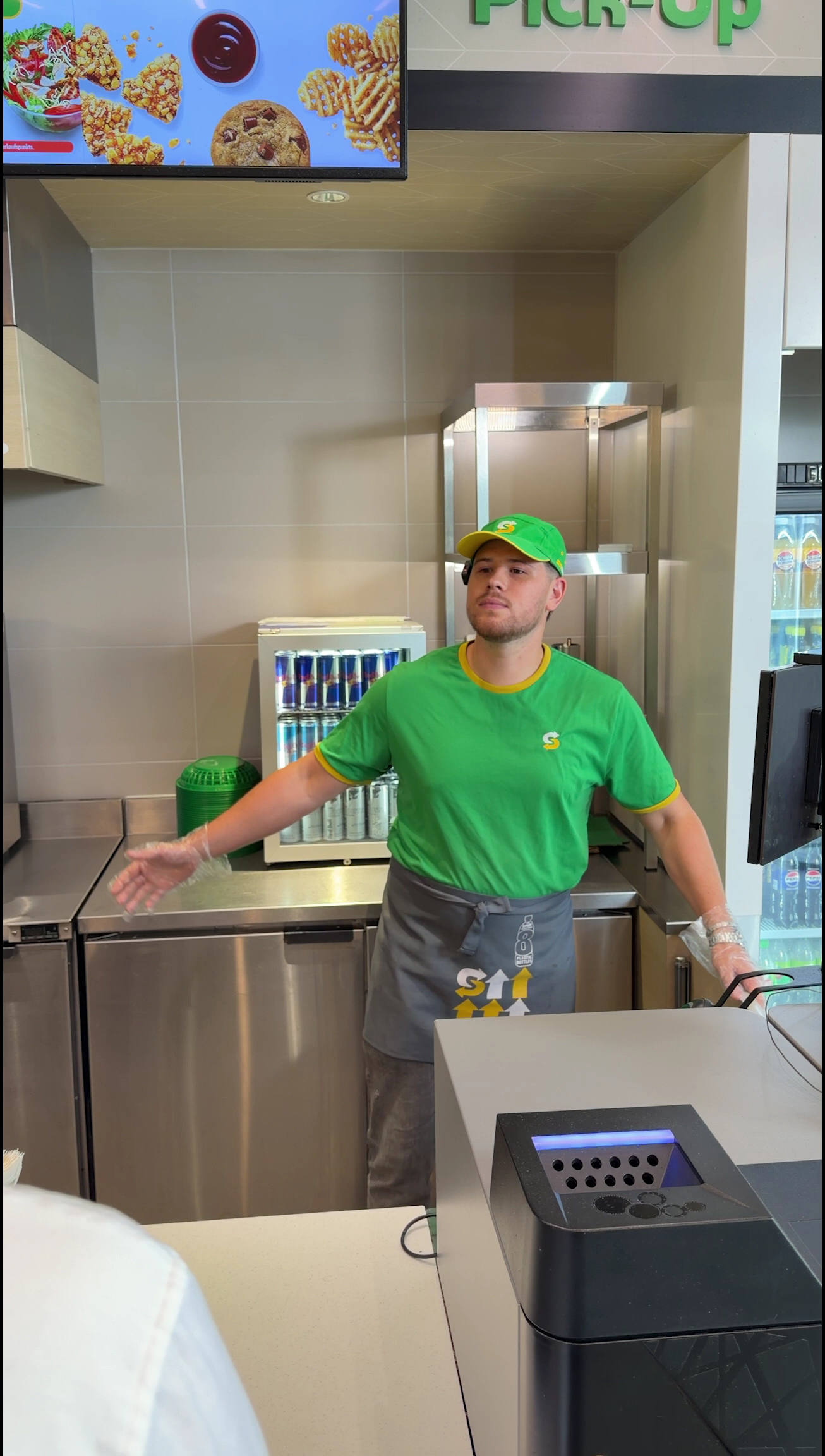 Subway employee in a green shirt and cap standing behind the restaurant counter in a campaign still.