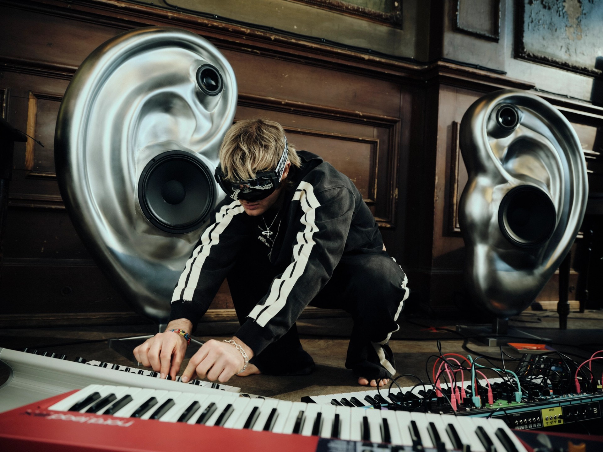 A performer crouches over a synthesizer between metallic ear-shaped speakers in the SkiAgguMusik campaign.