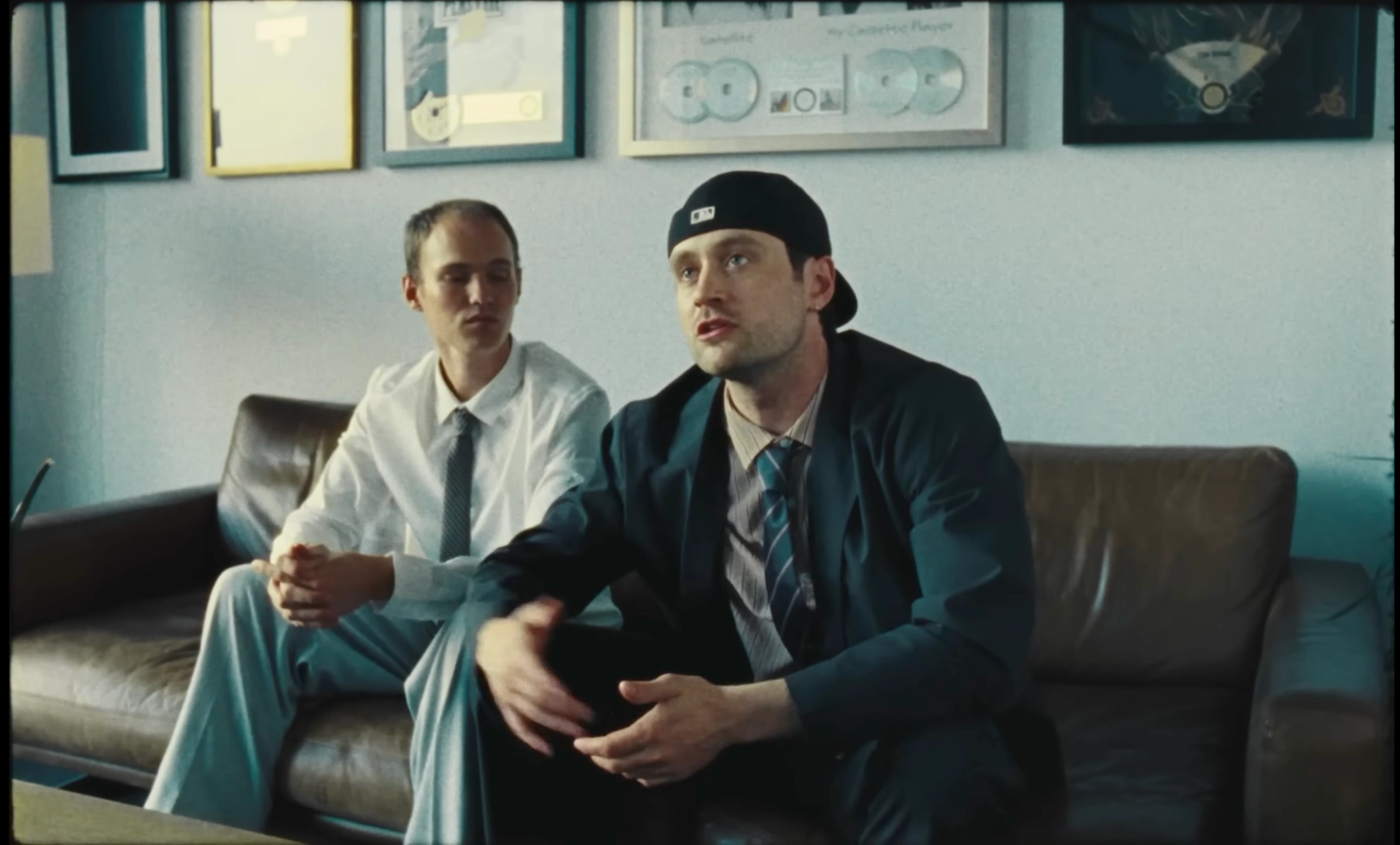Flaschko and Ritter Lean on the leather sofa: Flaschko in a white shirt and tie listens while Ritter Lean, in a backwards cap and dark suit, talks with his hands.