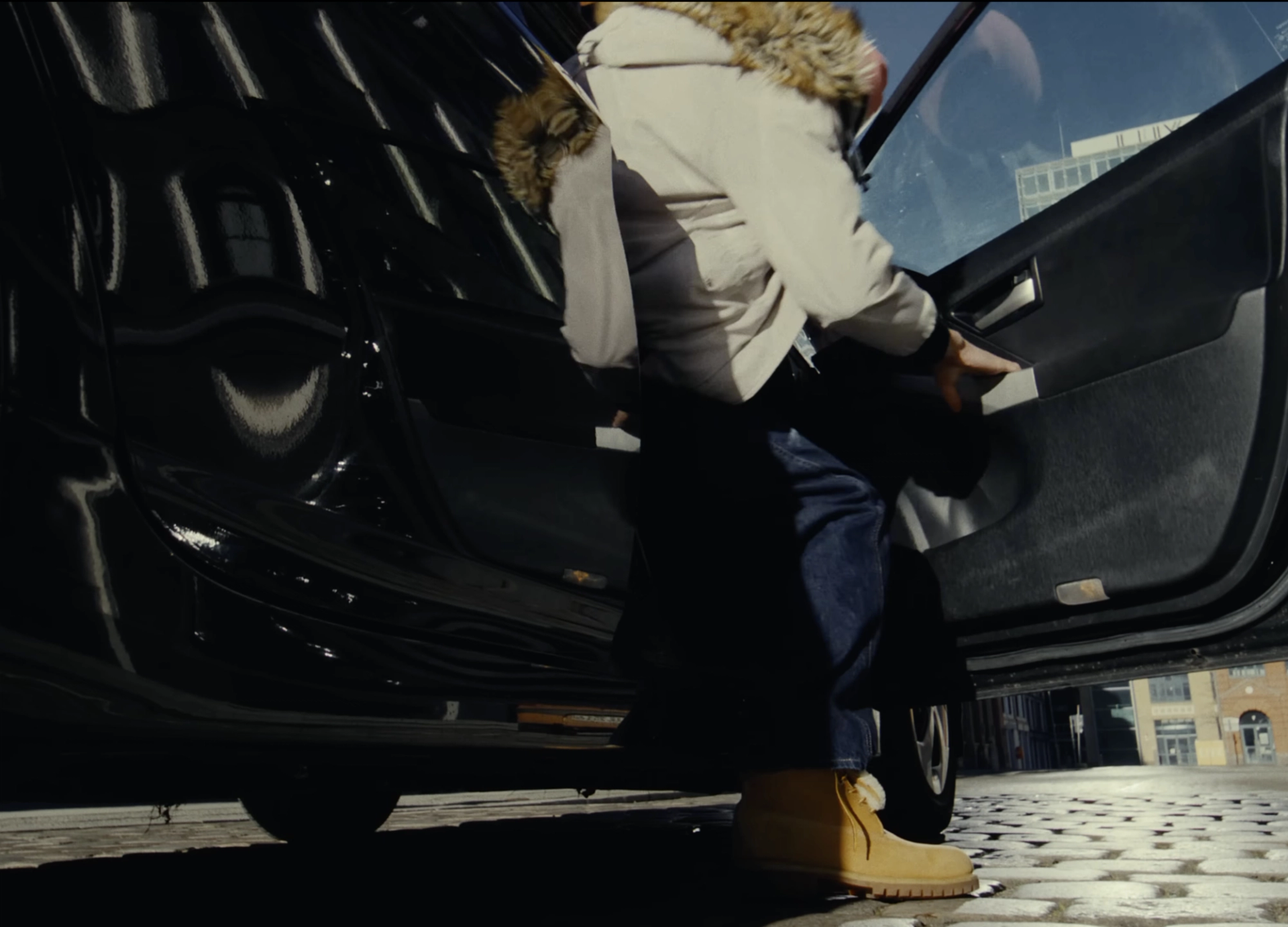 Low-angle shot of a person in a fur-hooded parka and tan boots climbing into a black car on a cobbled street.
