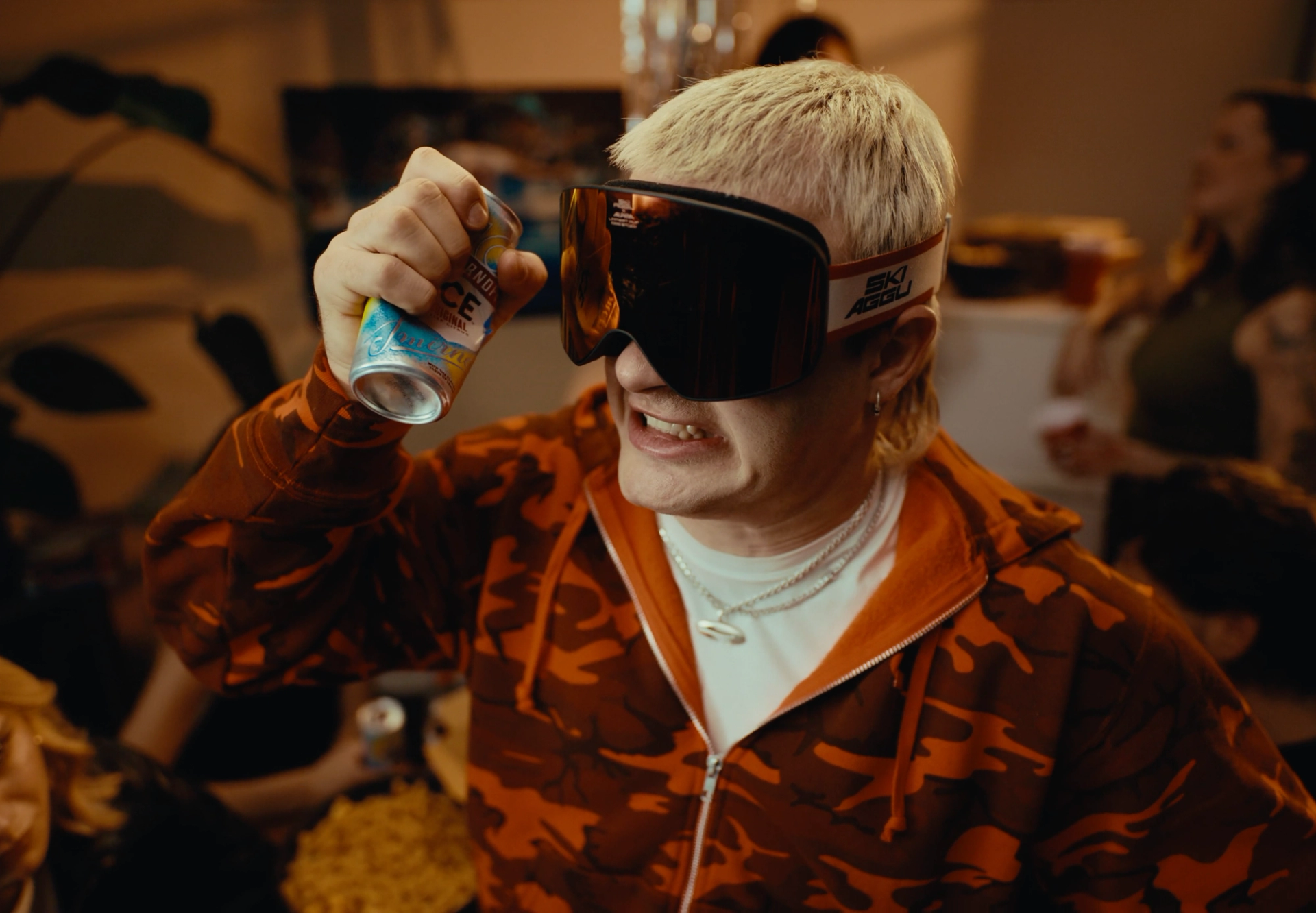 Close-up of Ski Aggu grimacing as he holds a Smirnoff Ice can up to his goggles, whose strap reads "SKI AGGU".