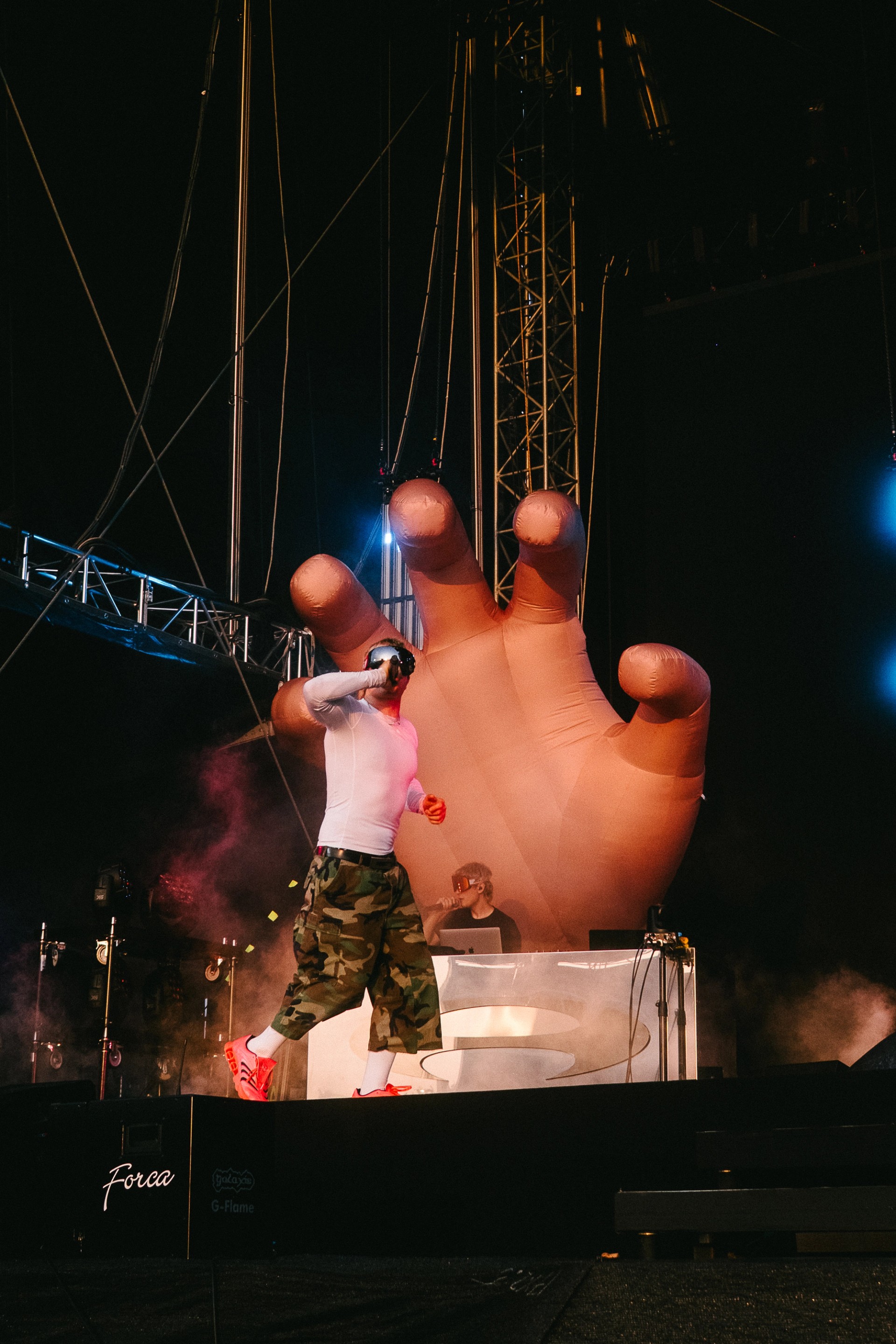 A performer in a white top and camouflage shorts strides across the stage in front of the giant inflatable hand, a DJ at a white booth behind him.