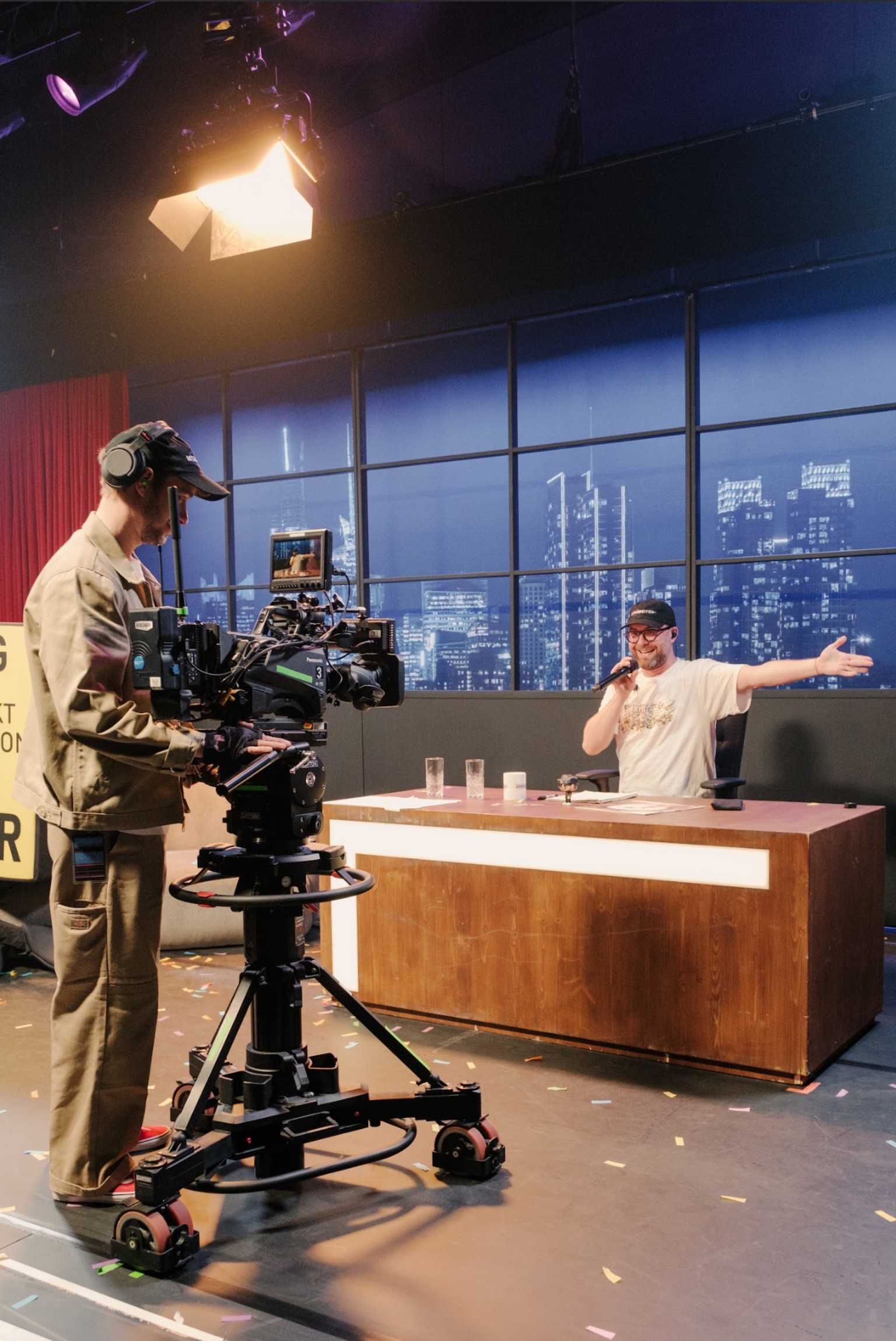 The late-night set on stage: the artist sits at the wooden host desk with a microphone, filmed by a TV camera operator, a night skyline behind him.