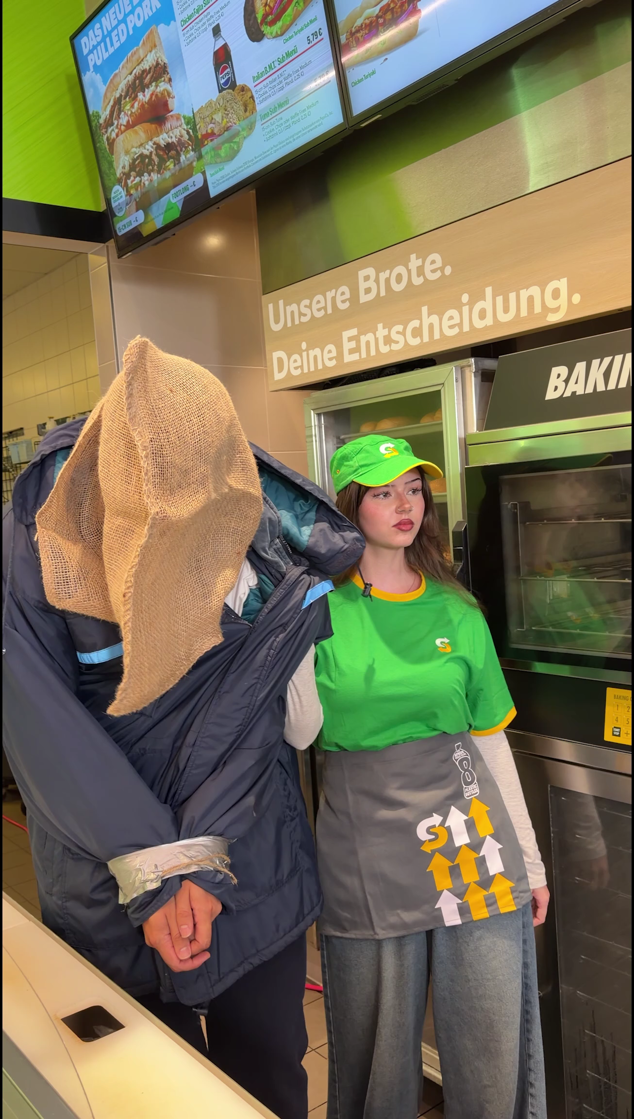 Subway campaign still of a hooded figure beside a member of staff in a green uniform at the counter.