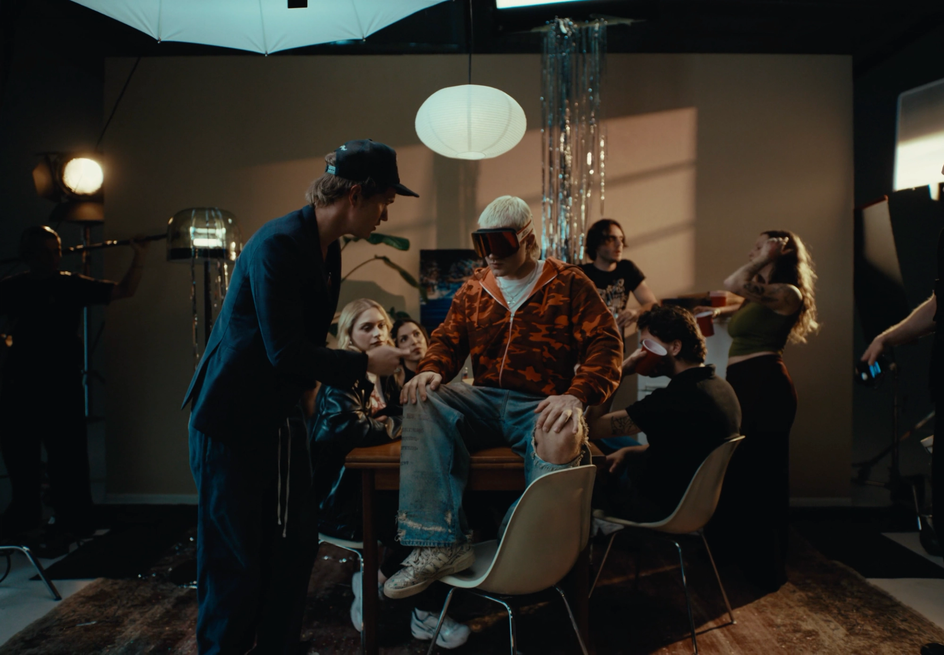 Party set seen from outside the scene: Flaschko in a cap talks to Ski Aggu, who sits on a table among the guests, with studio lamps at the edges.