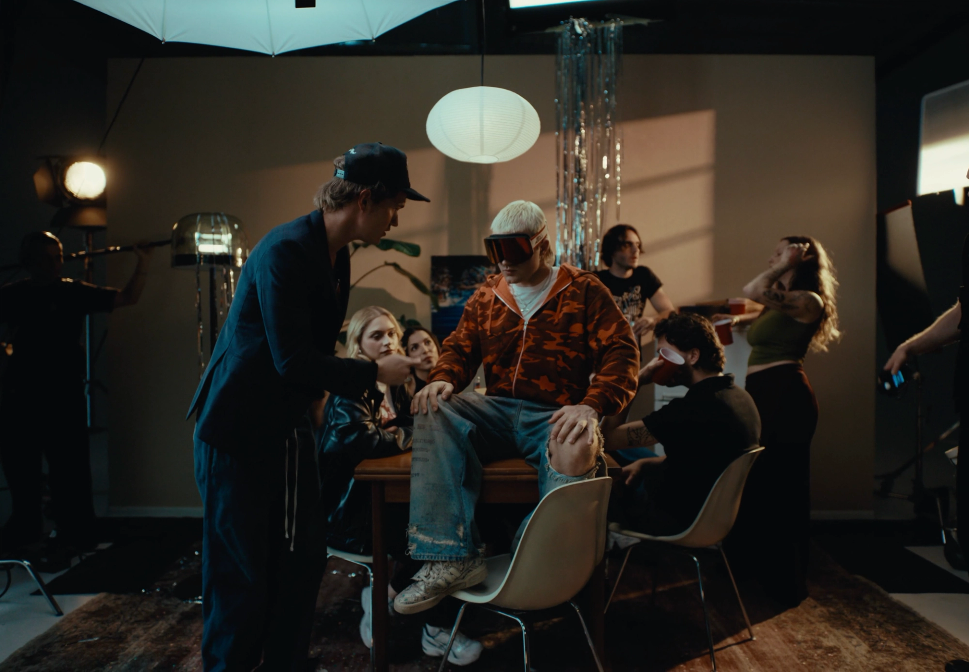 Party set seen from outside the scene: Flaschko in a cap talks to Ski Aggu, who sits on a table among the guests, with studio lamps at the edges.
