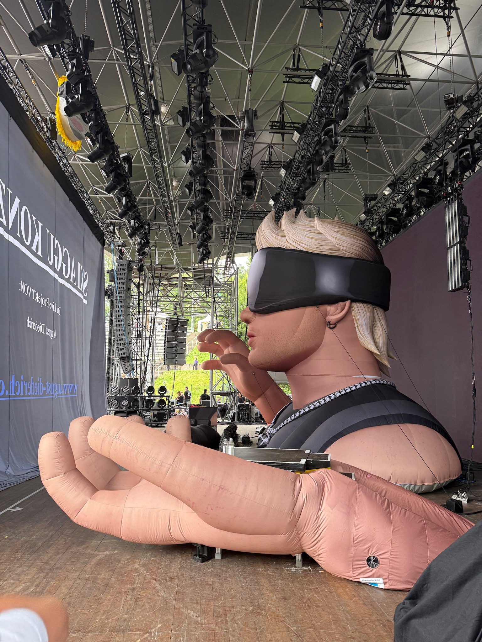 The inflatable in daylight during set-up, seen from the side: a blond head in black goggles with a chain, one giant hand lying flat on the stage floor.
