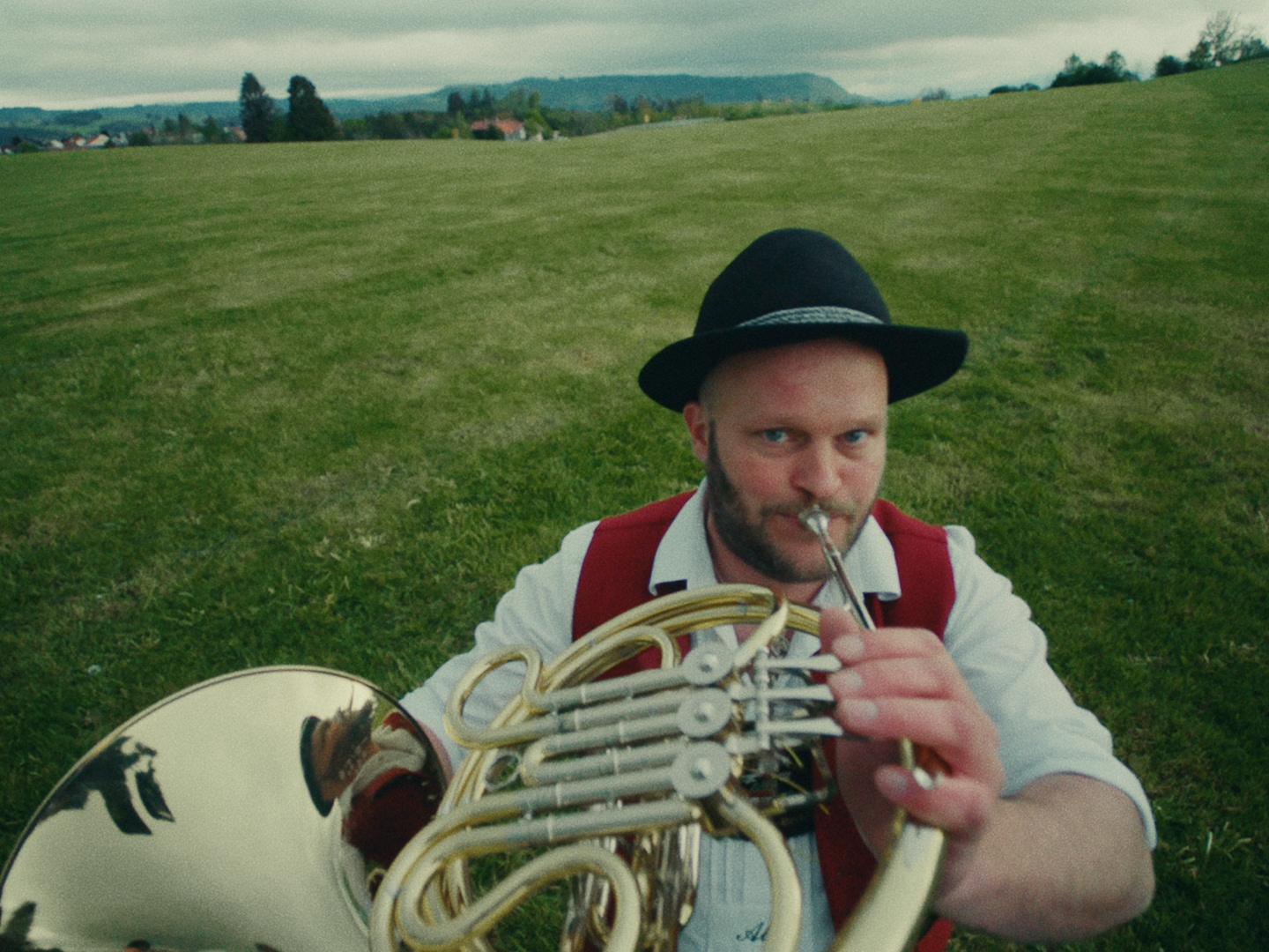 Horn player in a black hat and red waistcoat blows a French horn straight towards the camera in a green meadow.