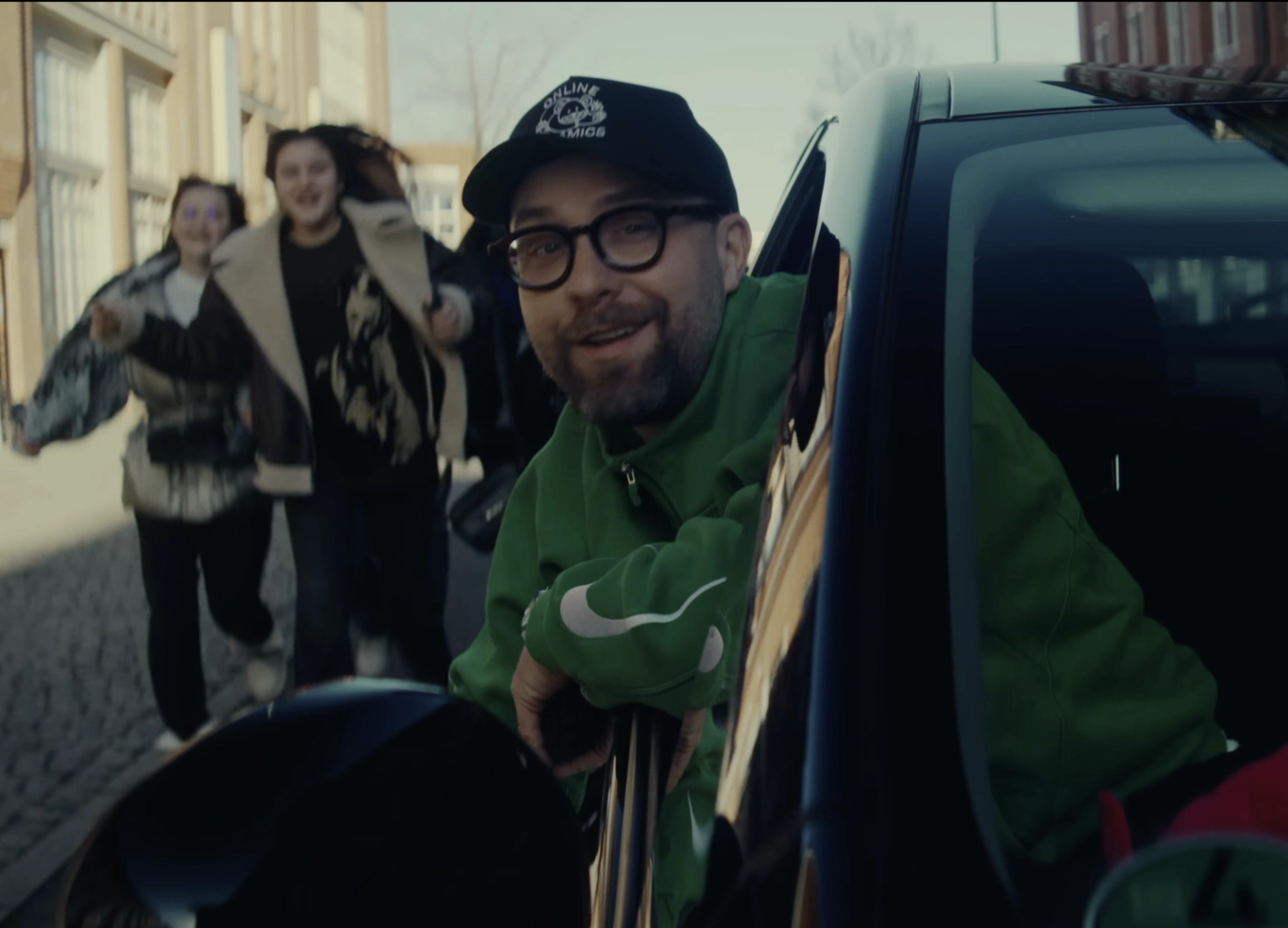 Leaning out of the window of the black car, Mark Forster in the green jacket and cap grins at the camera while two people run after it down the street.