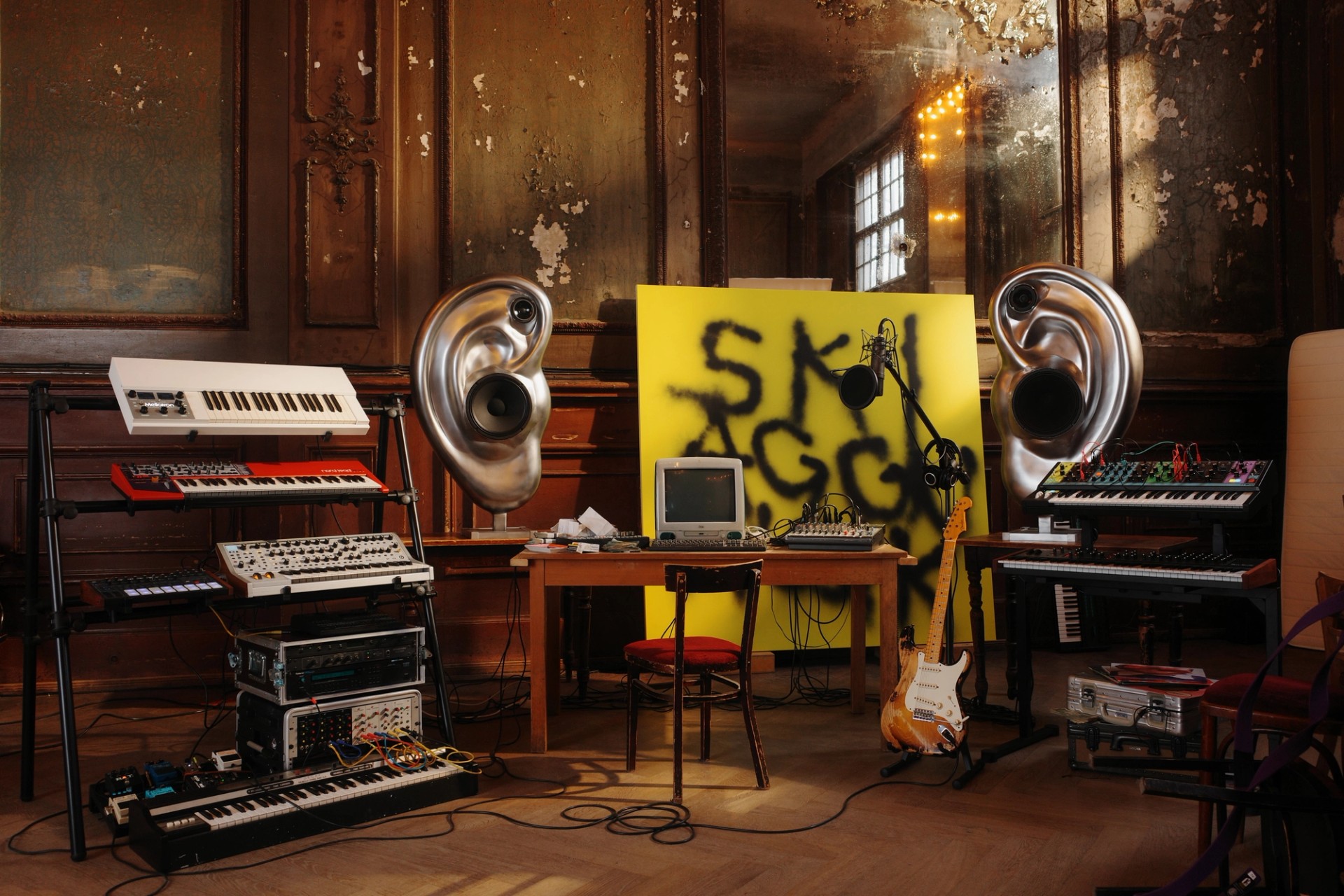 SkiAgguMusik set arranged around a desk, with stacked keyboards, a guitar and two metallic ear-shaped speakers.