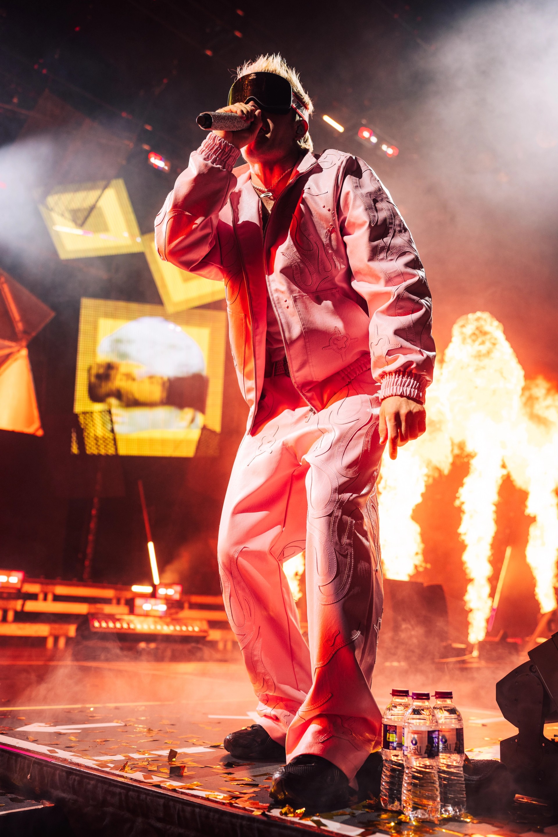 Flame jets fire behind Ski Aggu, who raps at the stage edge in a pale pink suit and ski goggles under yellow-lit LED panels hung at angles.