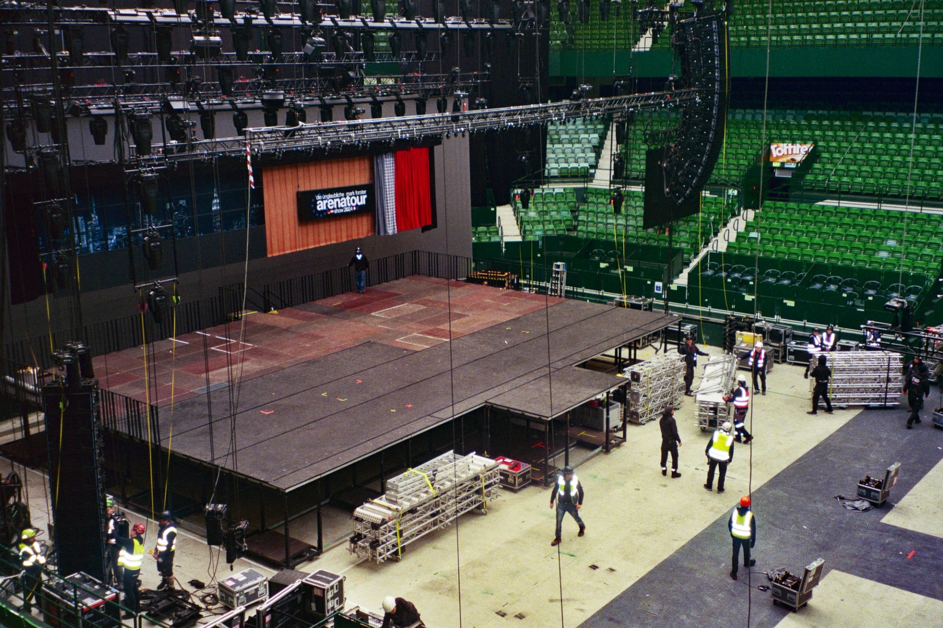 Mark Forster arena stage during assembly, with an exposed platform, overhead lighting rig and crew on the empty arena floor.