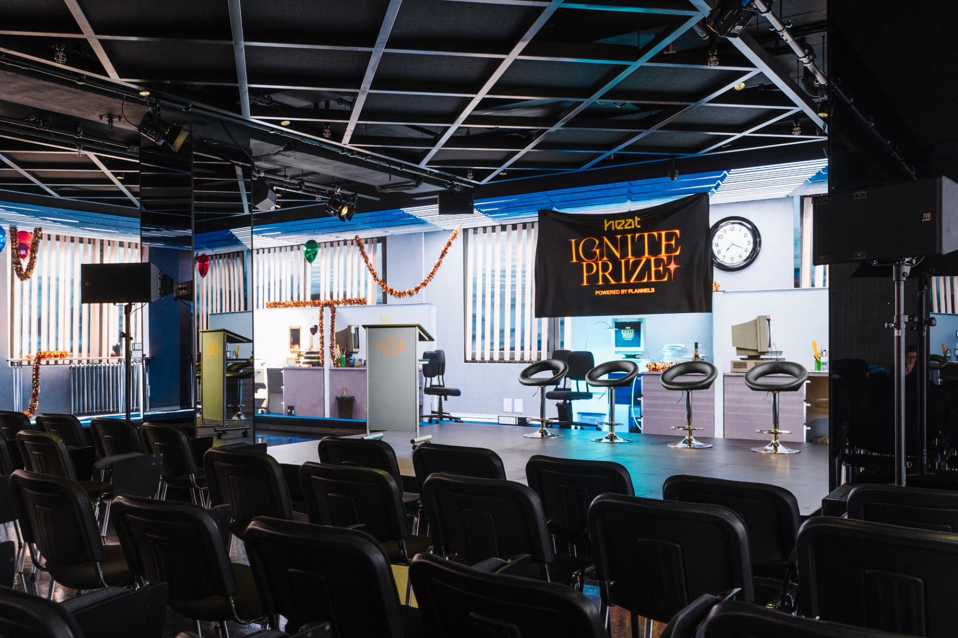 Event space with rows of black chairs facing a stage with four bar stools, a black "IGNITE PRIZE" banner and wall screens showing a tinsel-decorated 3D office.
