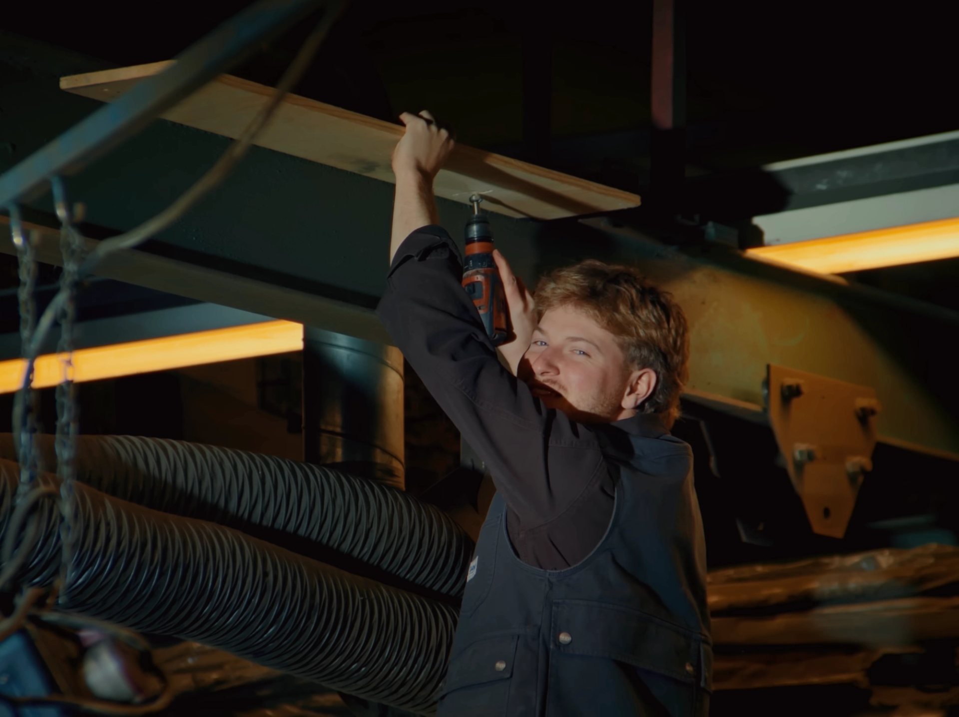 One of the band screws a wooden board to the workshop ceiling with a cordless drill and looks back over his shoulder at the camera.