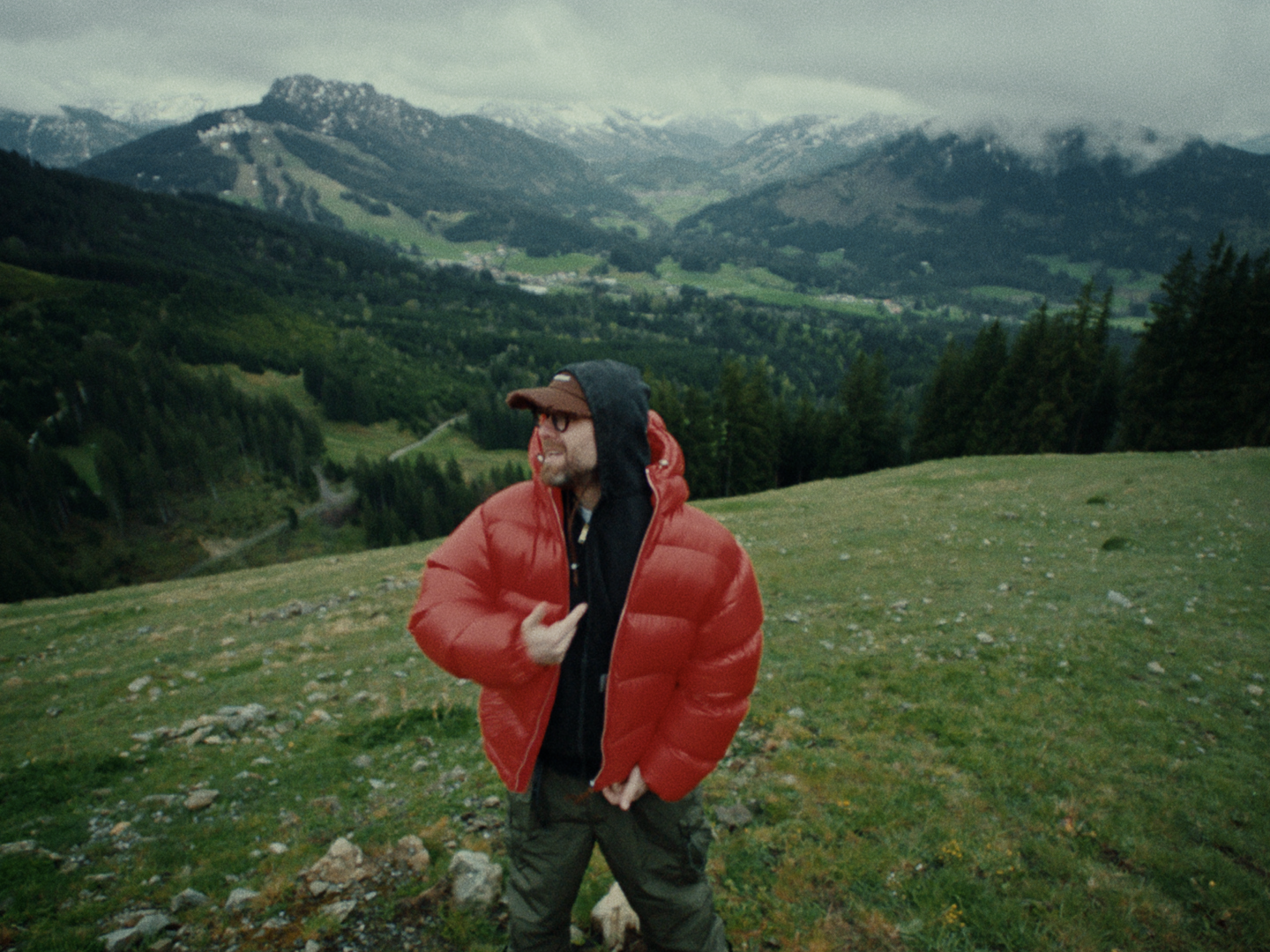 On an alpine meadow, Mark Forster in a red down jacket and cap looks to the side, with forested valleys and cloud-covered peaks behind him.