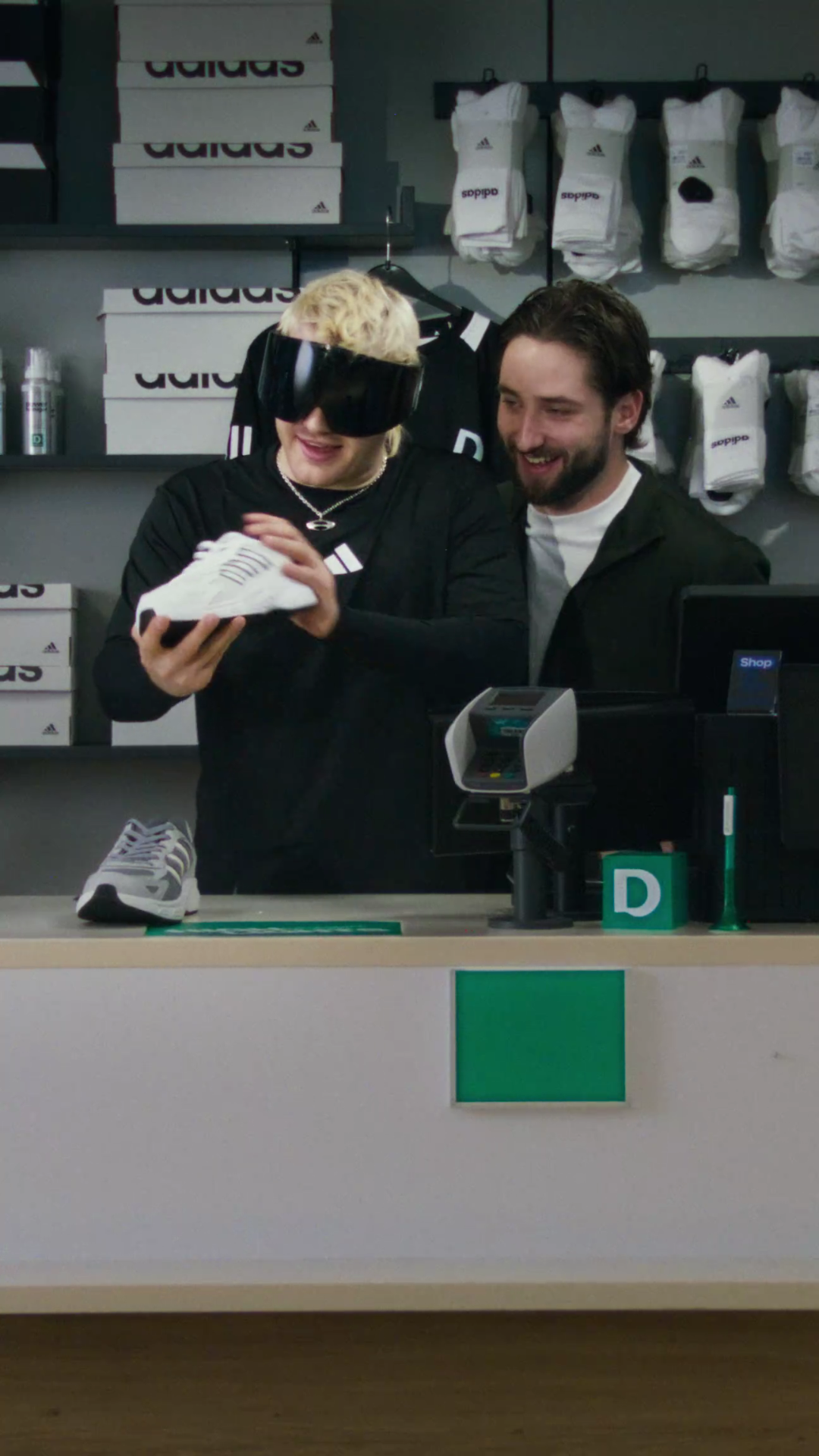 Behind a Deichmann shop counter, Ski Aggu in ski goggles turns a white Adidas sneaker in his hands while a bearded man looks on, shoe boxes and socks on the wall.