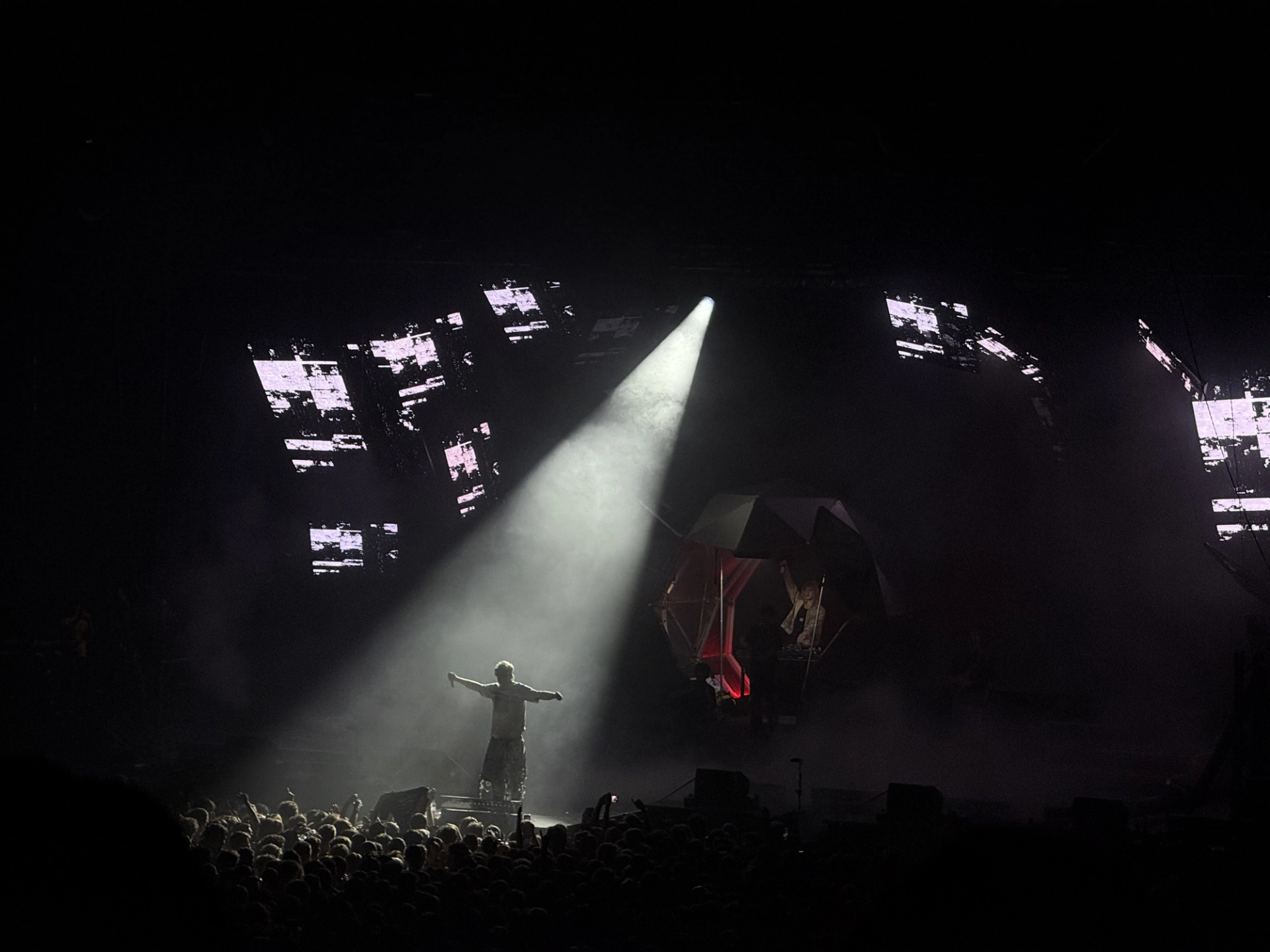 The stage from the back of a dark arena: one spotlight on Ski Aggu with arms spread, scattered LED screens around an angular, red-lit structure.
