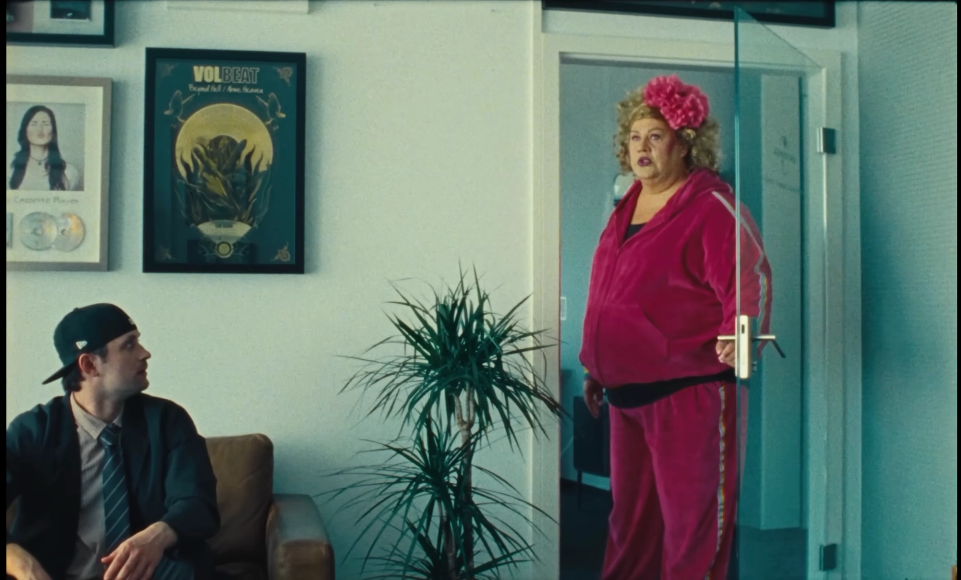 Cindy aus Marzahn, in a pink velour tracksuit with a pink flower in her hair, stands in a glass doorway while Ritter Lean looks up from the sofa.