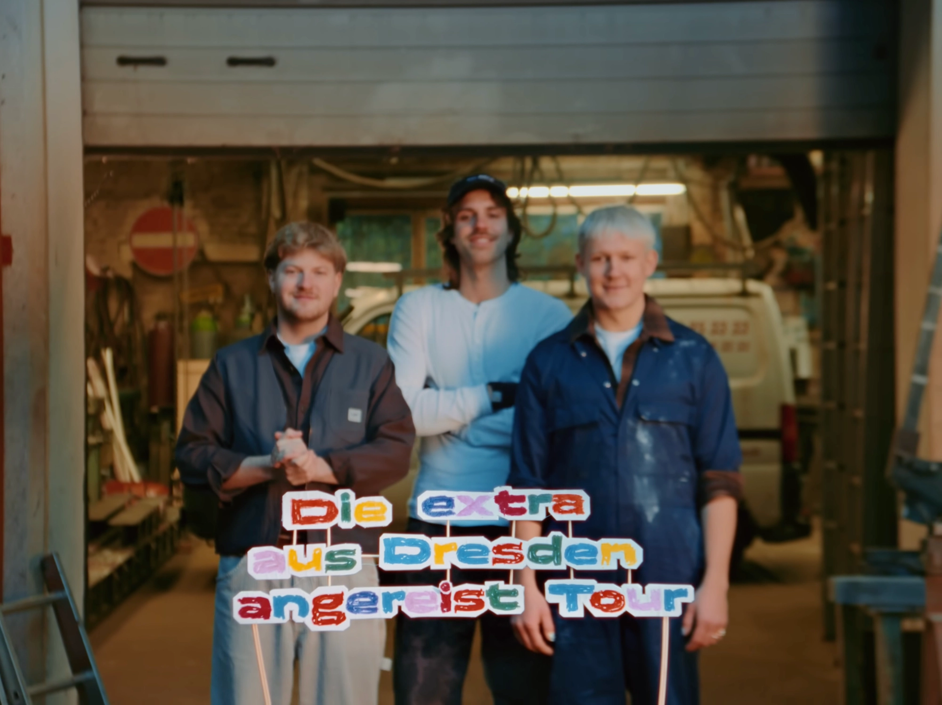 Hand-coloured sign reading "Die extra aus Dresden angereist Tour" in focus, the three band members standing blurred in the workshop gate behind it.