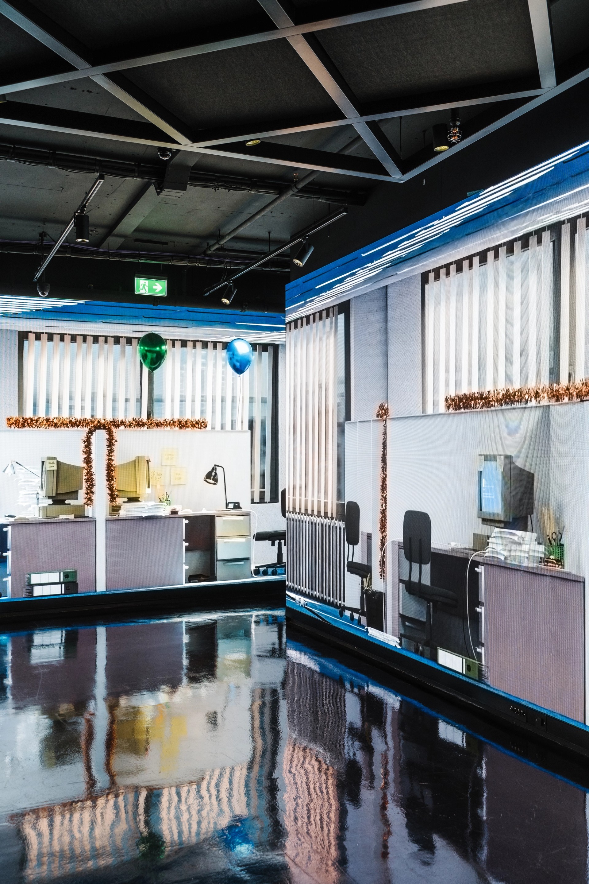 Upright view of two screens meeting at a corner, the rendered office desks, blinds and balloons mirrored in the glossy black floor.