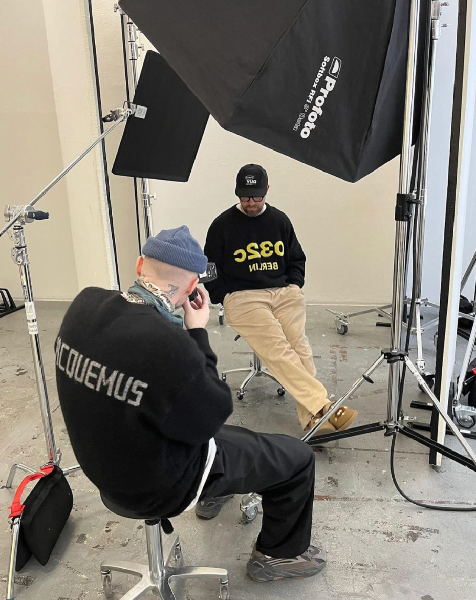Behind the scenes of a photo shoot: a photographer in a blue beanie, seen from behind, shoots Mark Forster seated on a stool under a large softbox.