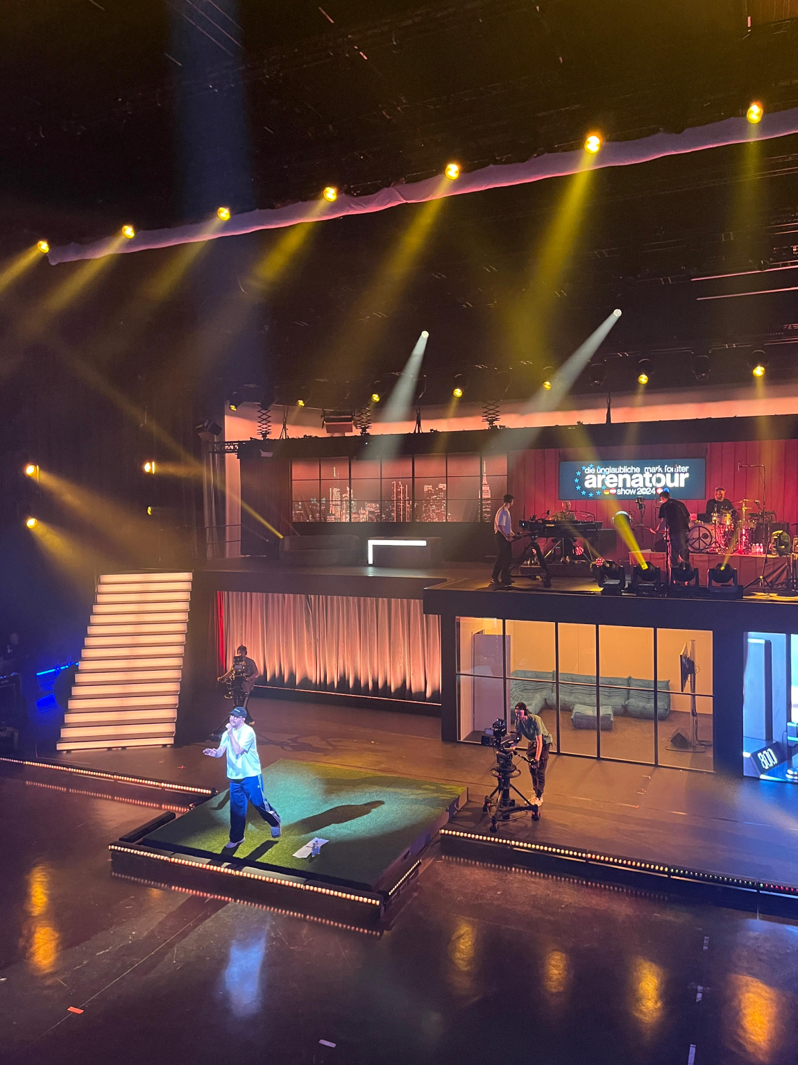 Soundcheck on the arena tour stage: a performer on the green platform, camera operators beside him and the band on the upper level.