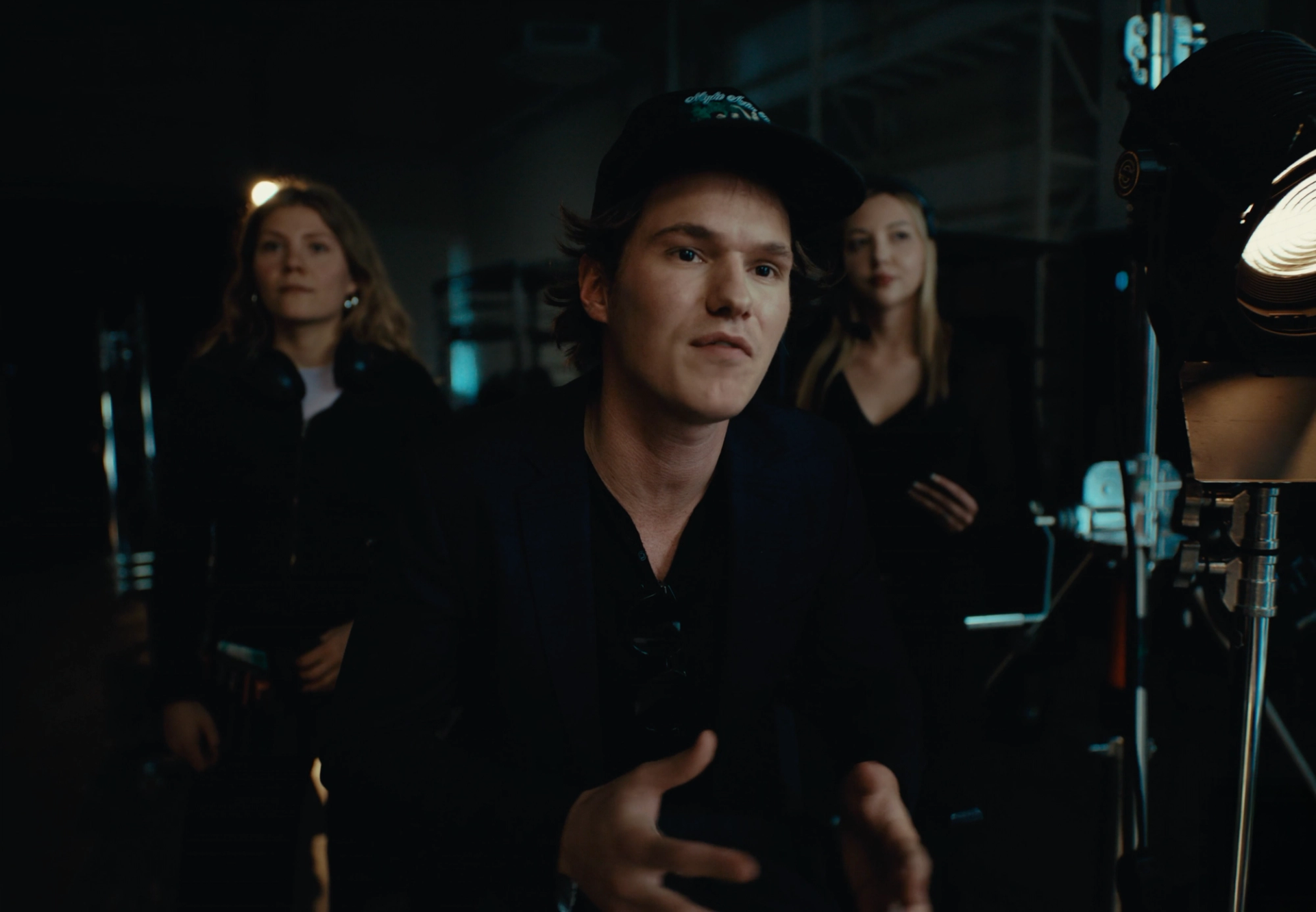 Still from the commercial: Flaschko, in the middle, explains something with open hands on the dark set, two women watching from behind.