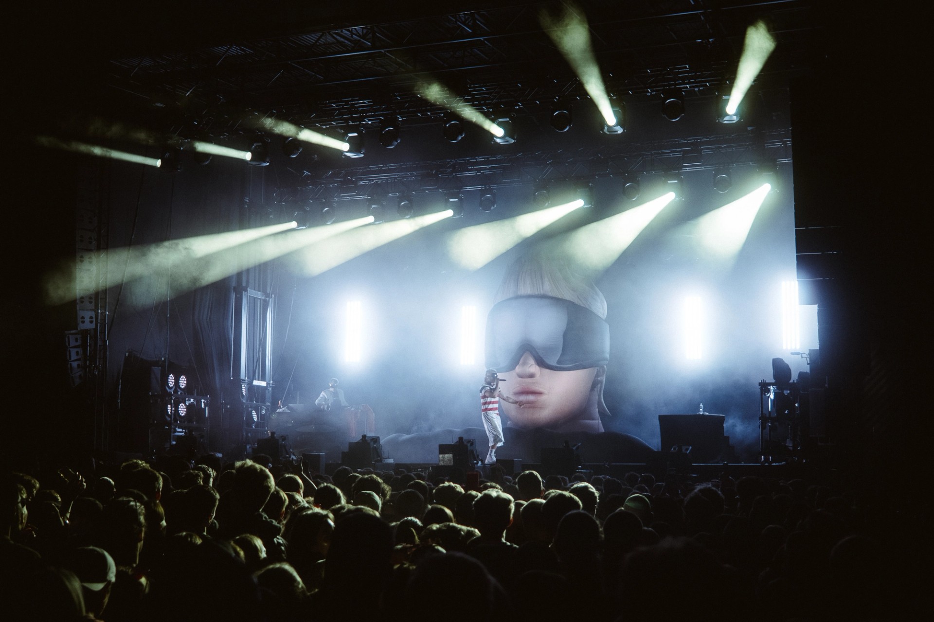 Wide view from the back of the crowd: the inflatable head in goggles glows behind a performer under white beams of light, the audience in silhouette.
